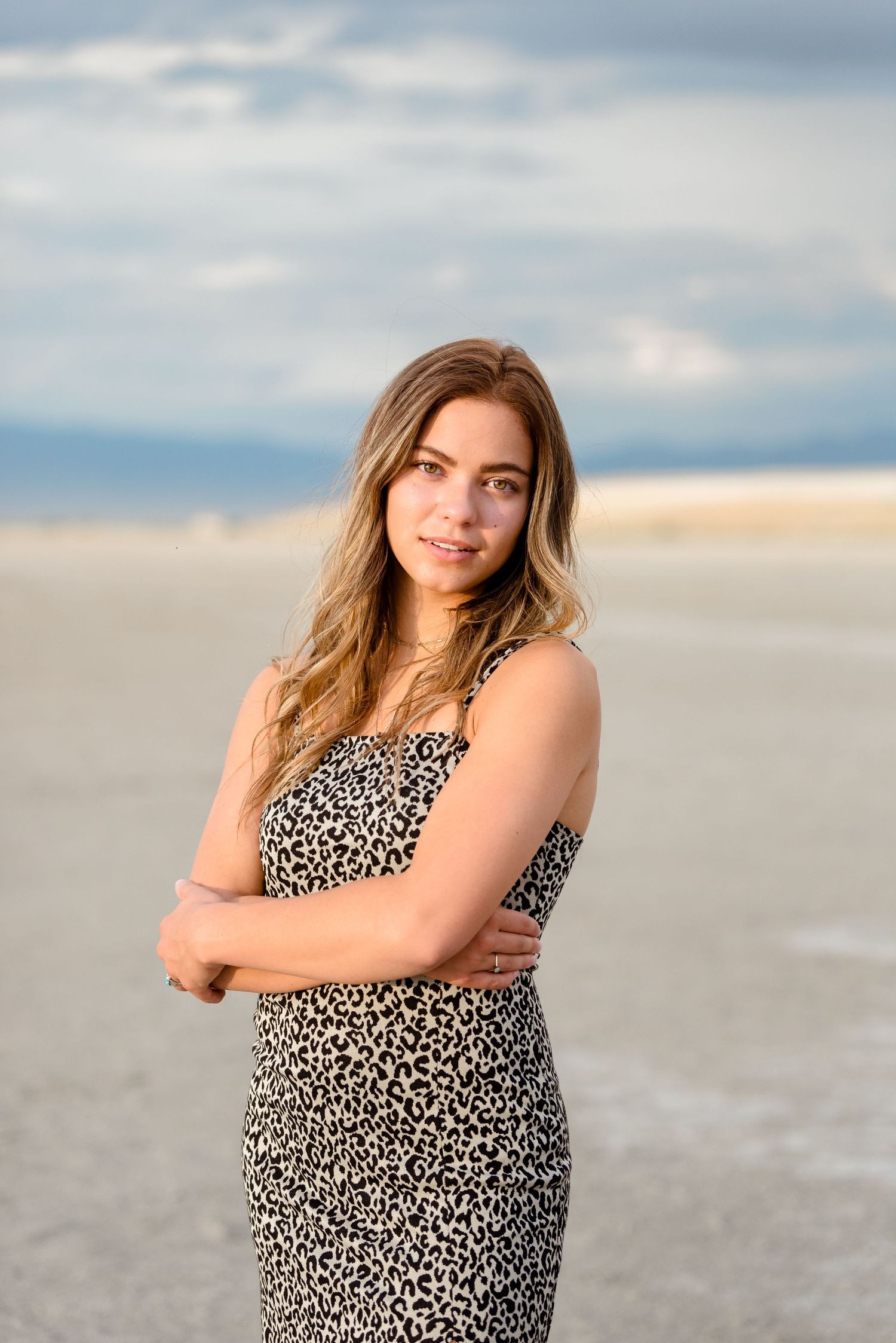 A person wearing a leopard print dress poses on a sandy beach during sunset with cloudy skies in the background.