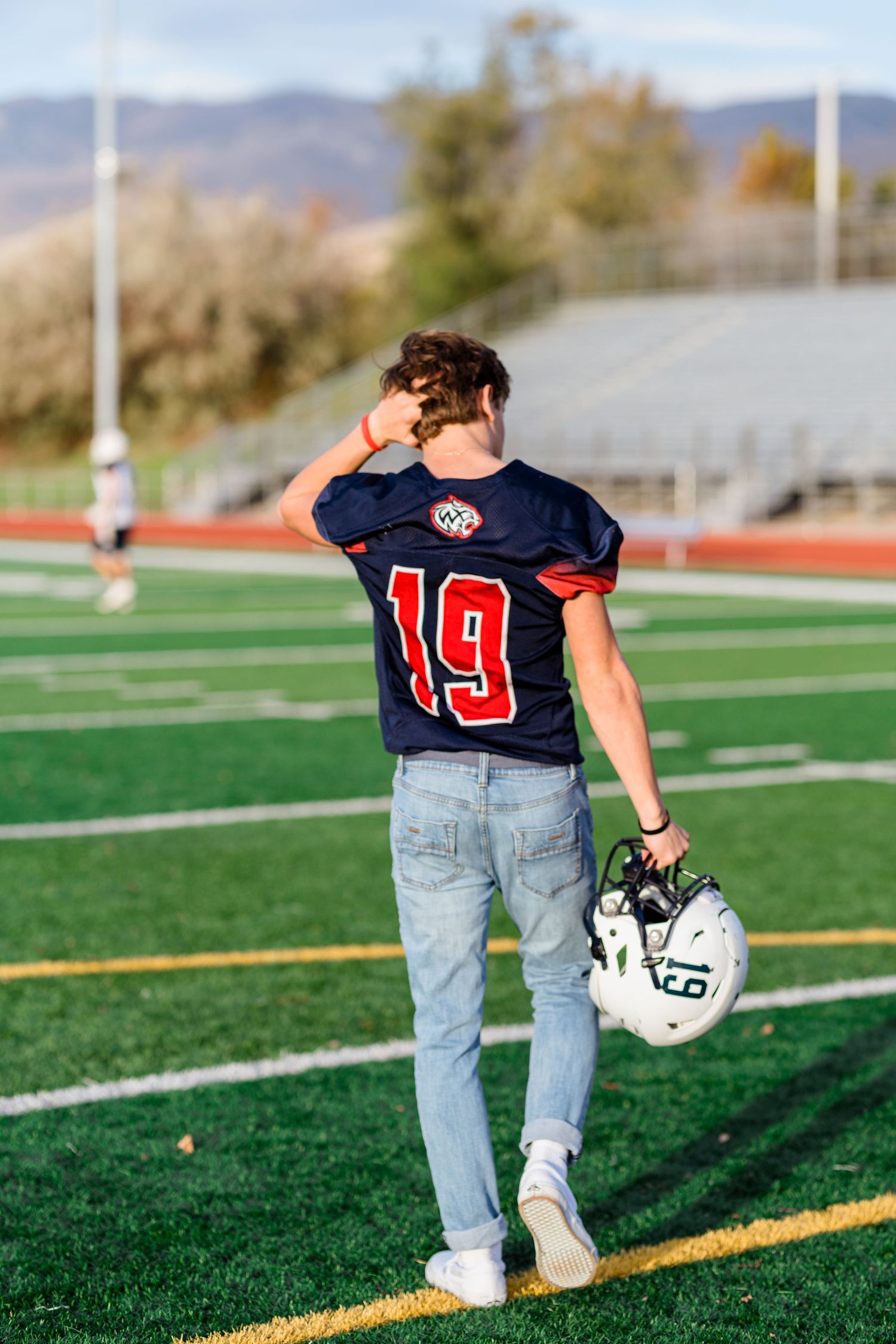 Football player in navy jersey number 19 walking across artificial turf field with helmet and mountains in background.