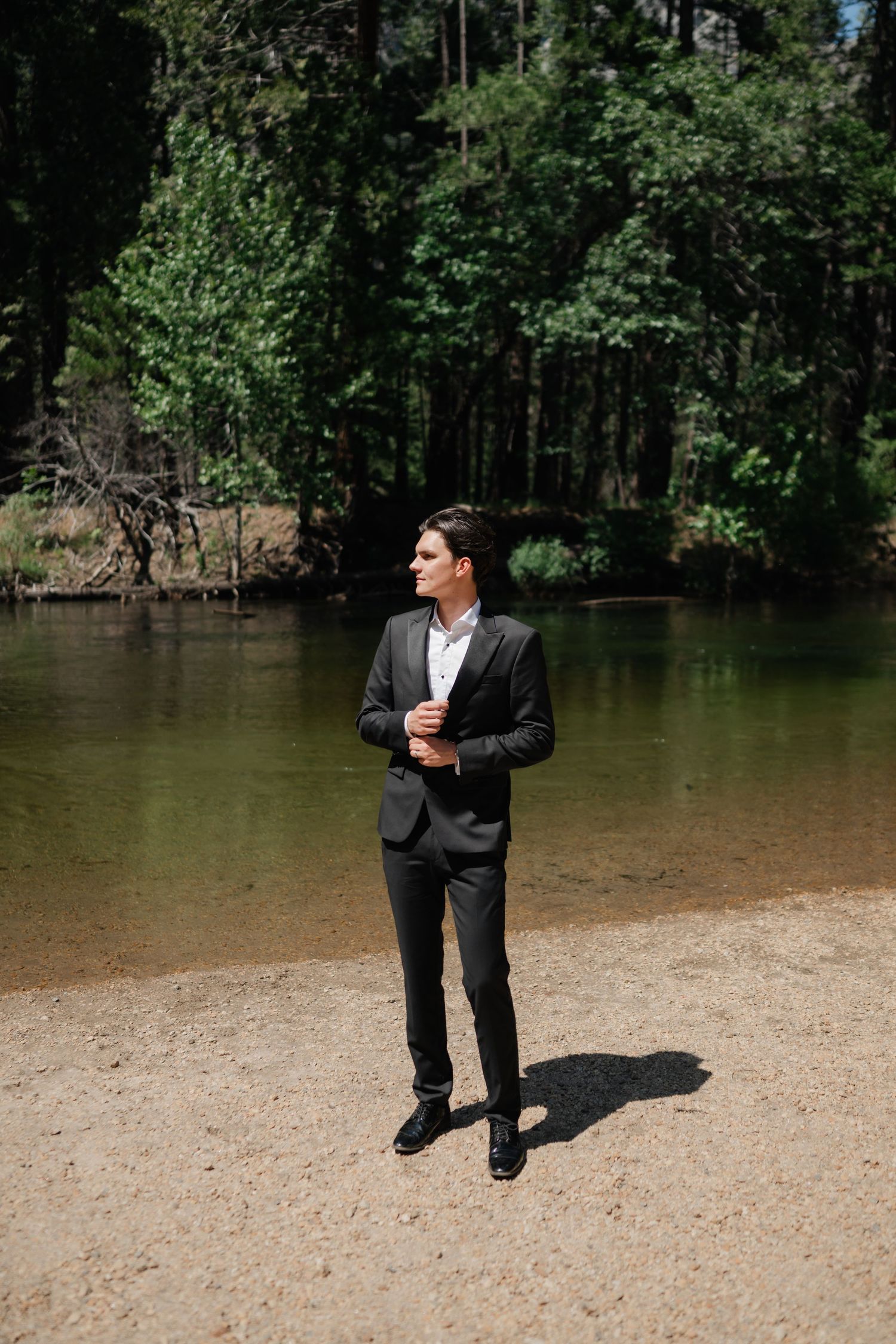 A well-dressed man in a dark suit stands on a sandy riverbank surrounded by lush green forest trees.