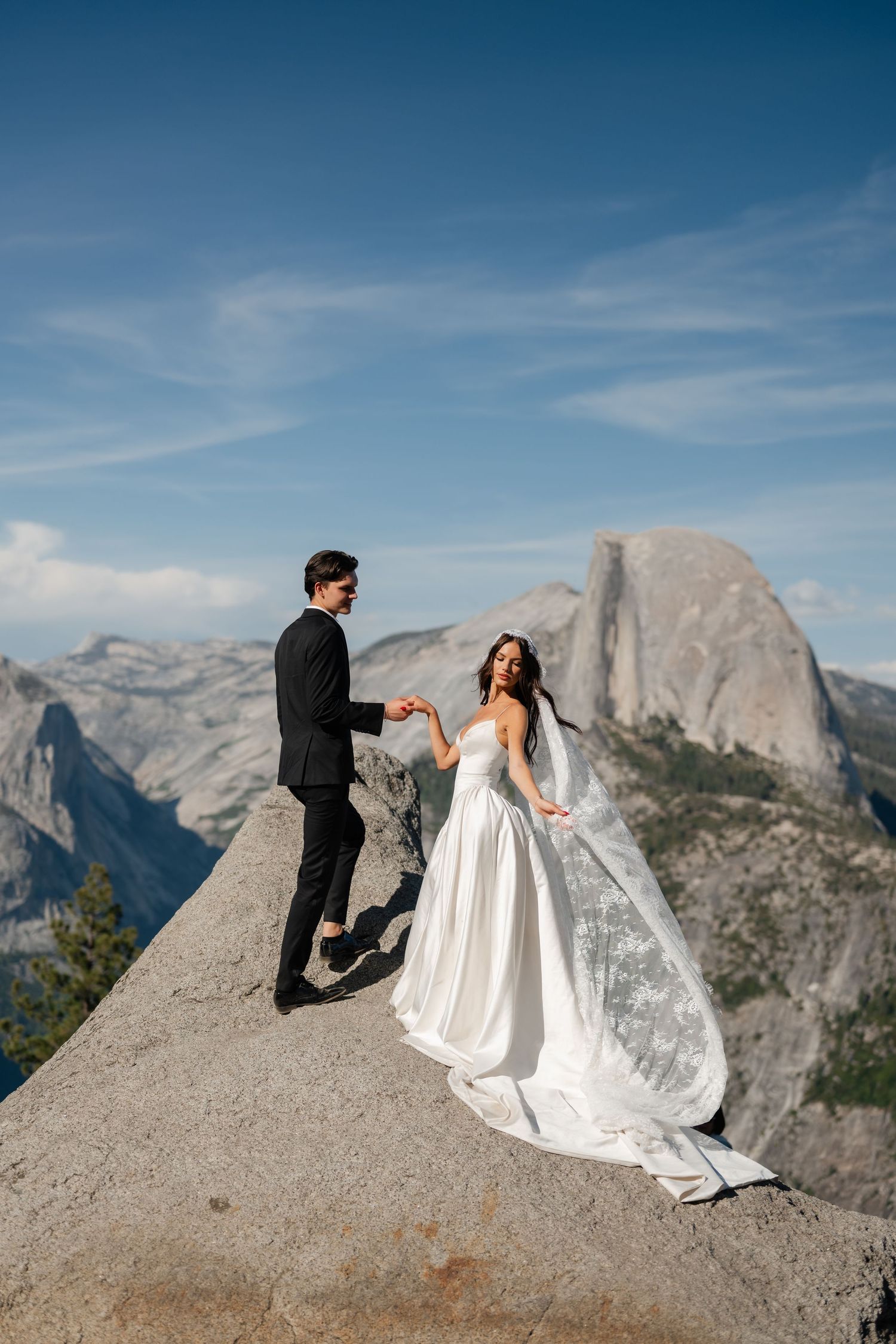Bride and groom hold hands on a rocky ledge at Yosemite with Half Dome in the background on their wedding day.