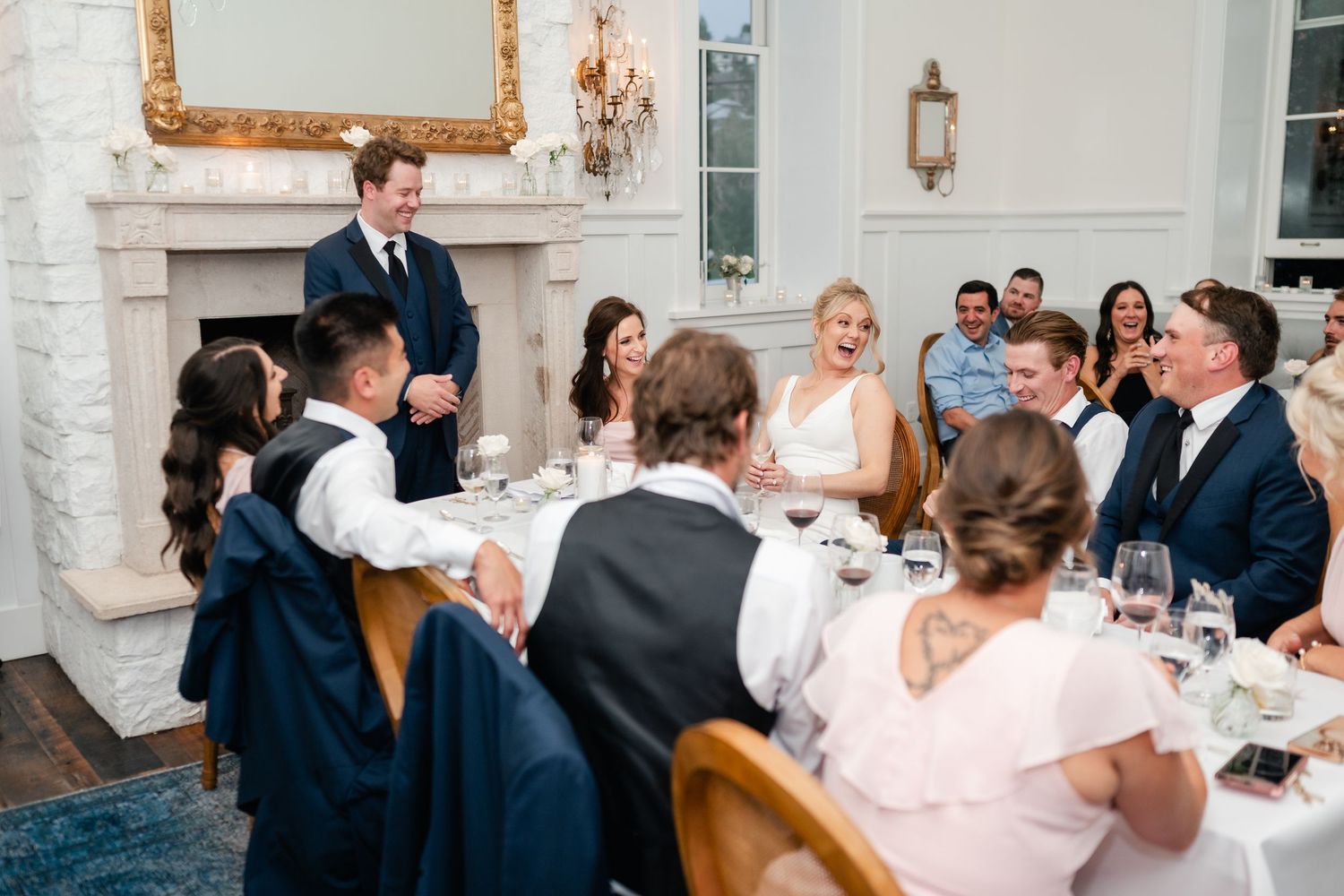 Guests laughing and celebrating at an elegant wedding reception dinner around a round table in a white decorated venue.