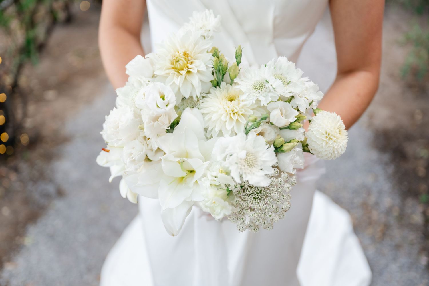 Bride holding a lush white wedding bouquet featuring dahlias, peonies, and baby's breath with a white ribbon.
