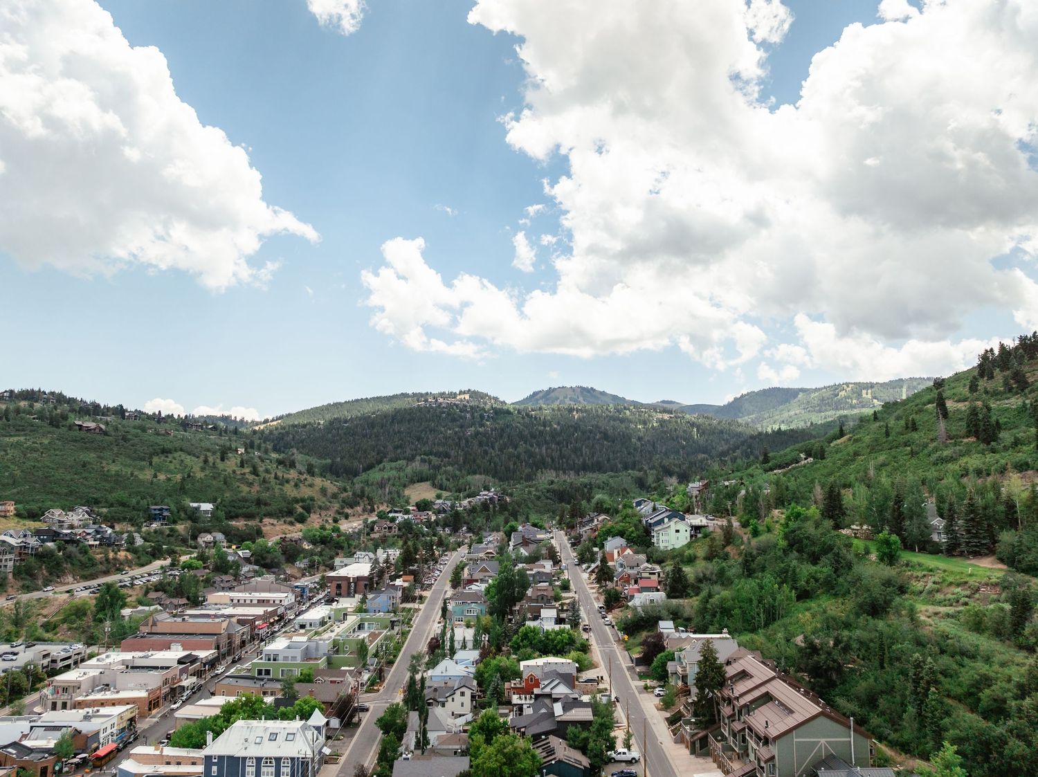 Aerial view of a Park City, Utah with tree-covered mountains, historic buildings, and historic main street under a partly cloudy sky.