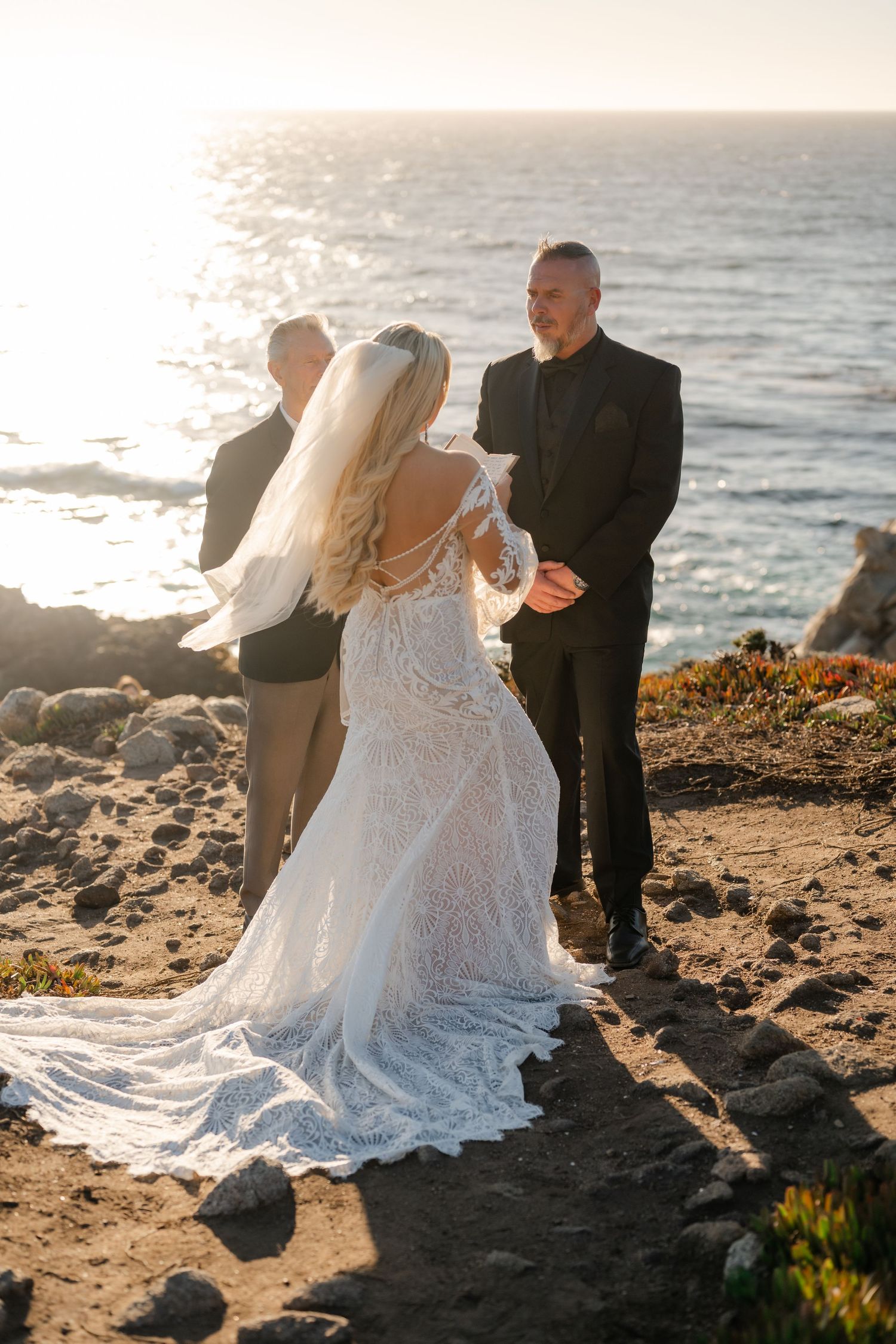 A romantic wedding ceremony takes place on a rocky coastal cliff at sunset with ocean views.