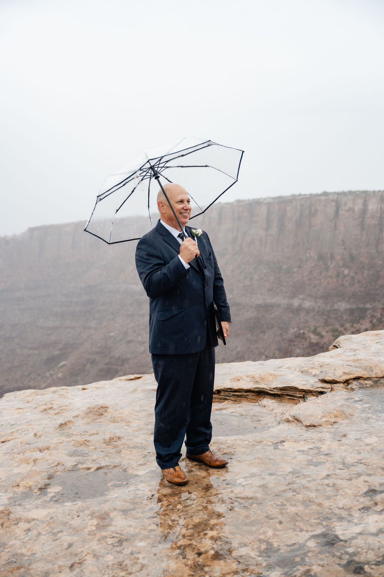 Person in navy suit holding clear umbrella while standing on canyon overlook.
