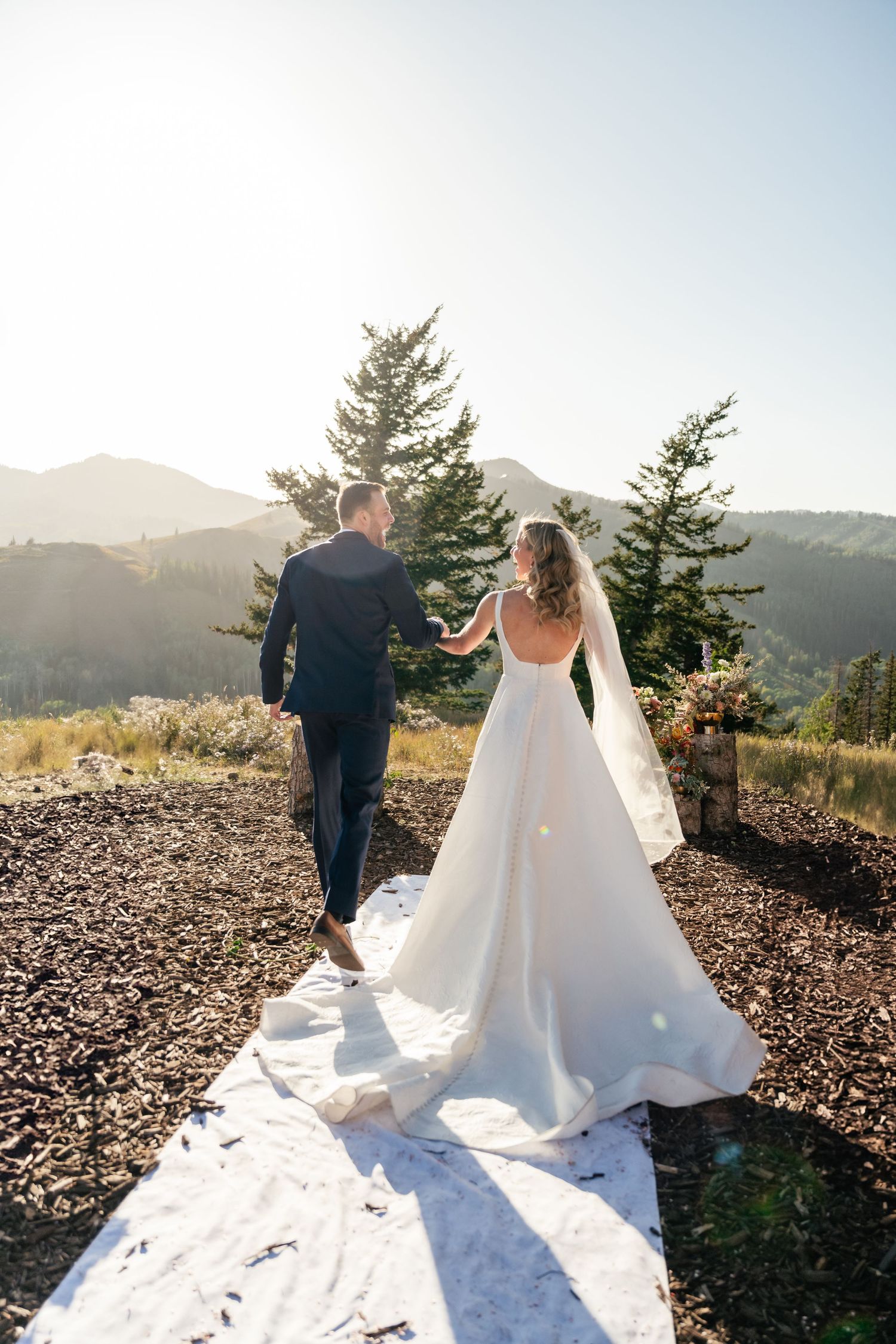Couple in wedding attire standing on rocky mountain overlook during sunset.