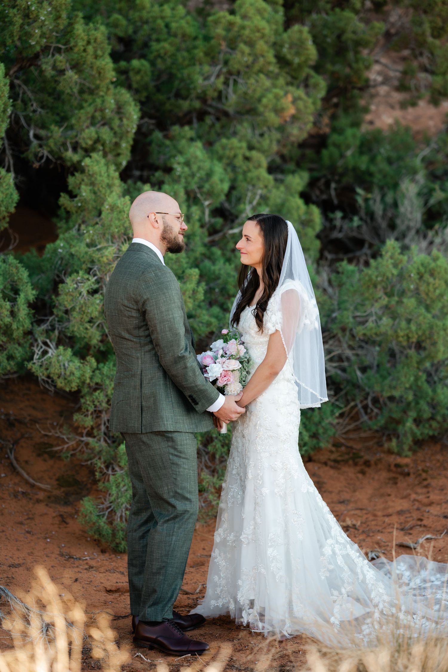 Wedding couple among desert juniper trees with red rocks in background.