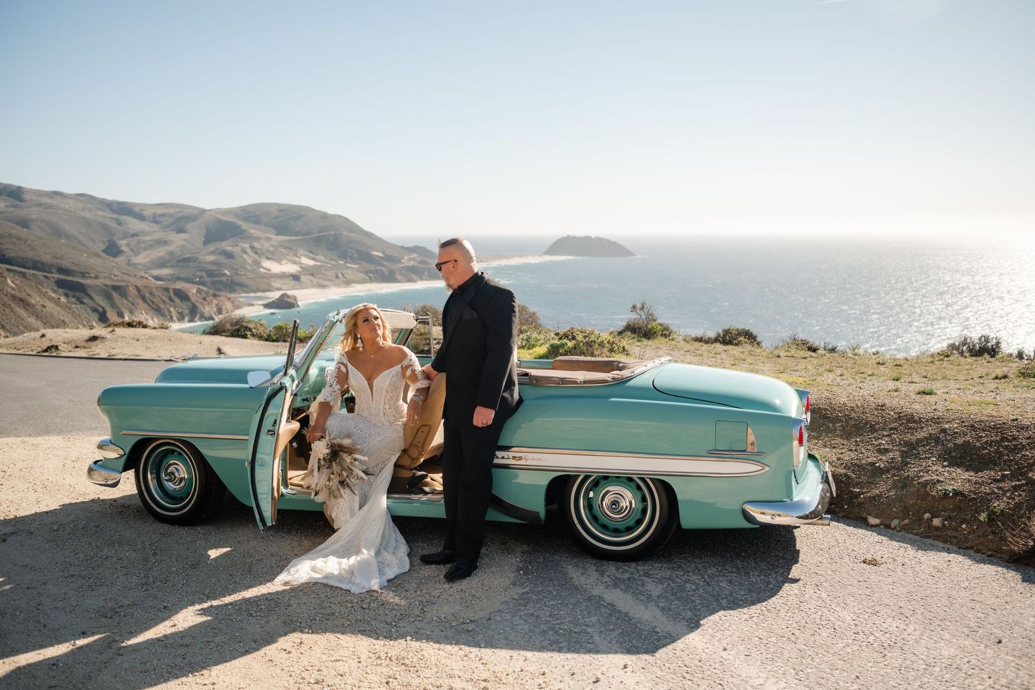 Vintage turquoise car against coastal backdrop with mountains and ocean.