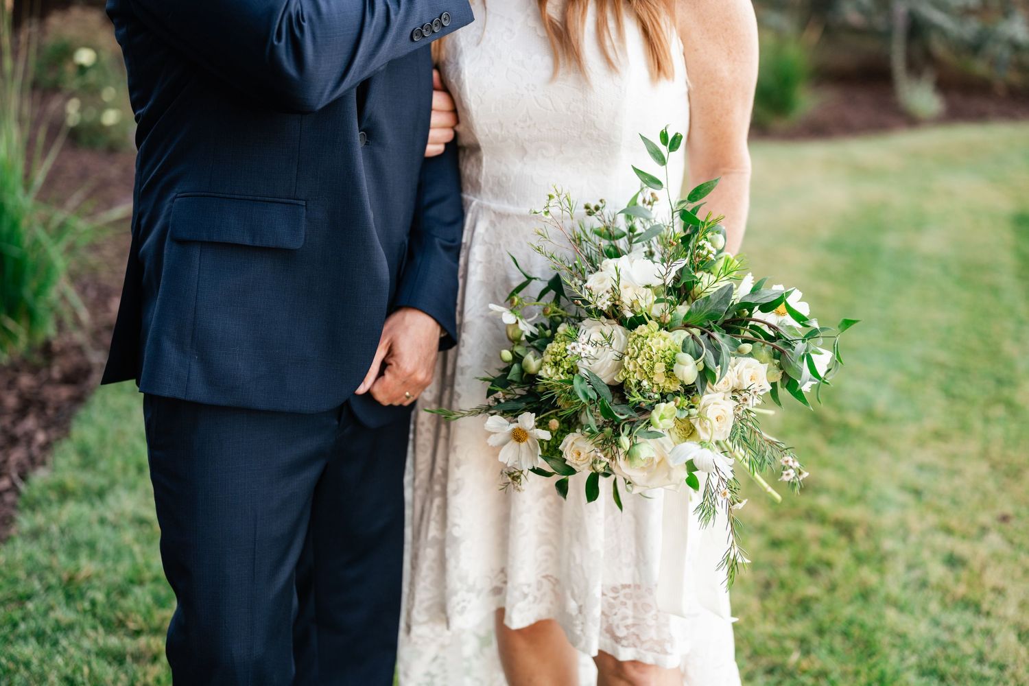 A couple stands close together on a grassy lawn during a wedding celebration with white floral bouquet details.
