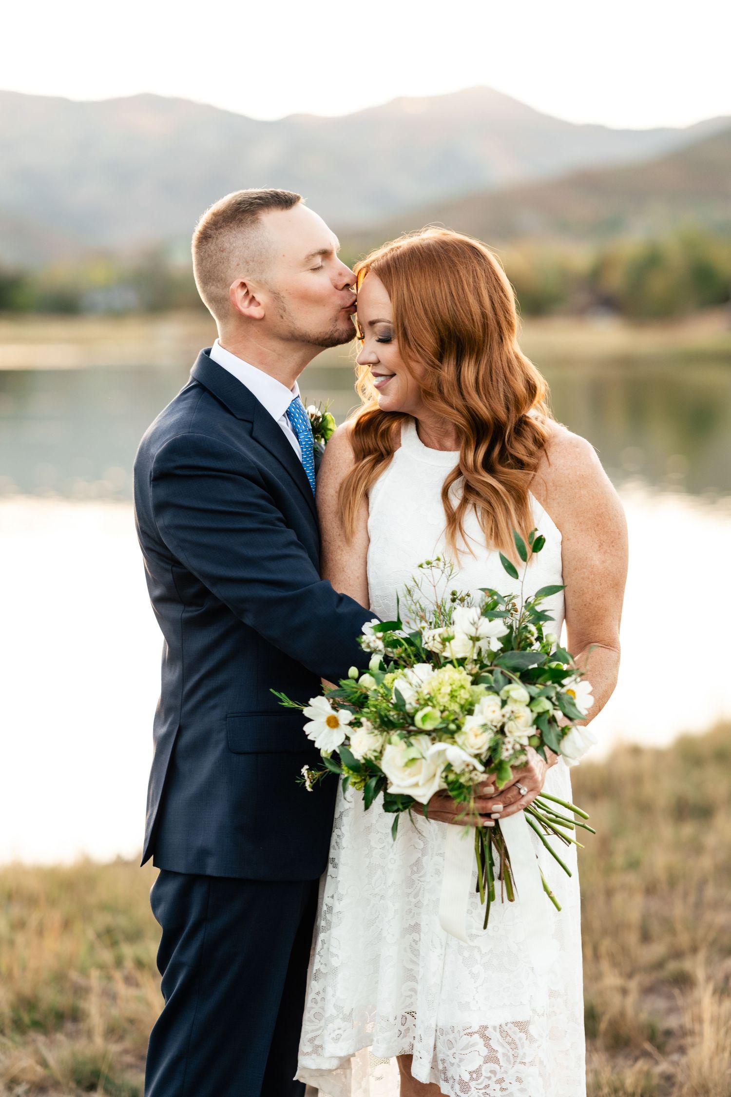 A romantic wedding couple shares intimate moments by a serene lake with white floral bouquet details. Park City Wedding Photographer.