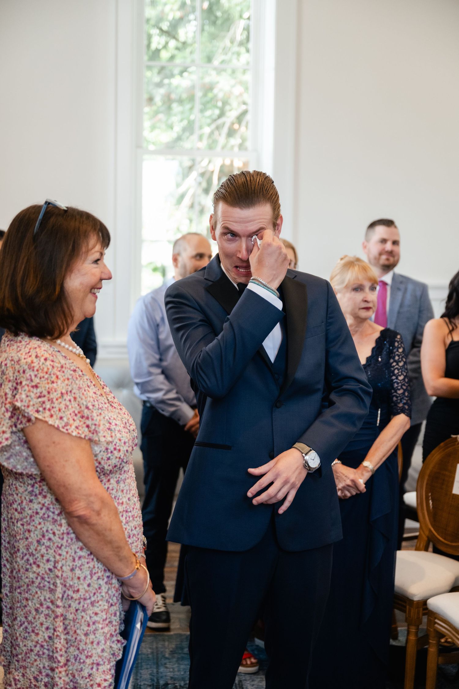 Emotional groom wiping tears at wedding ceremony, wearing navy suit, surrounded by guests in the Washington School House Hotel.