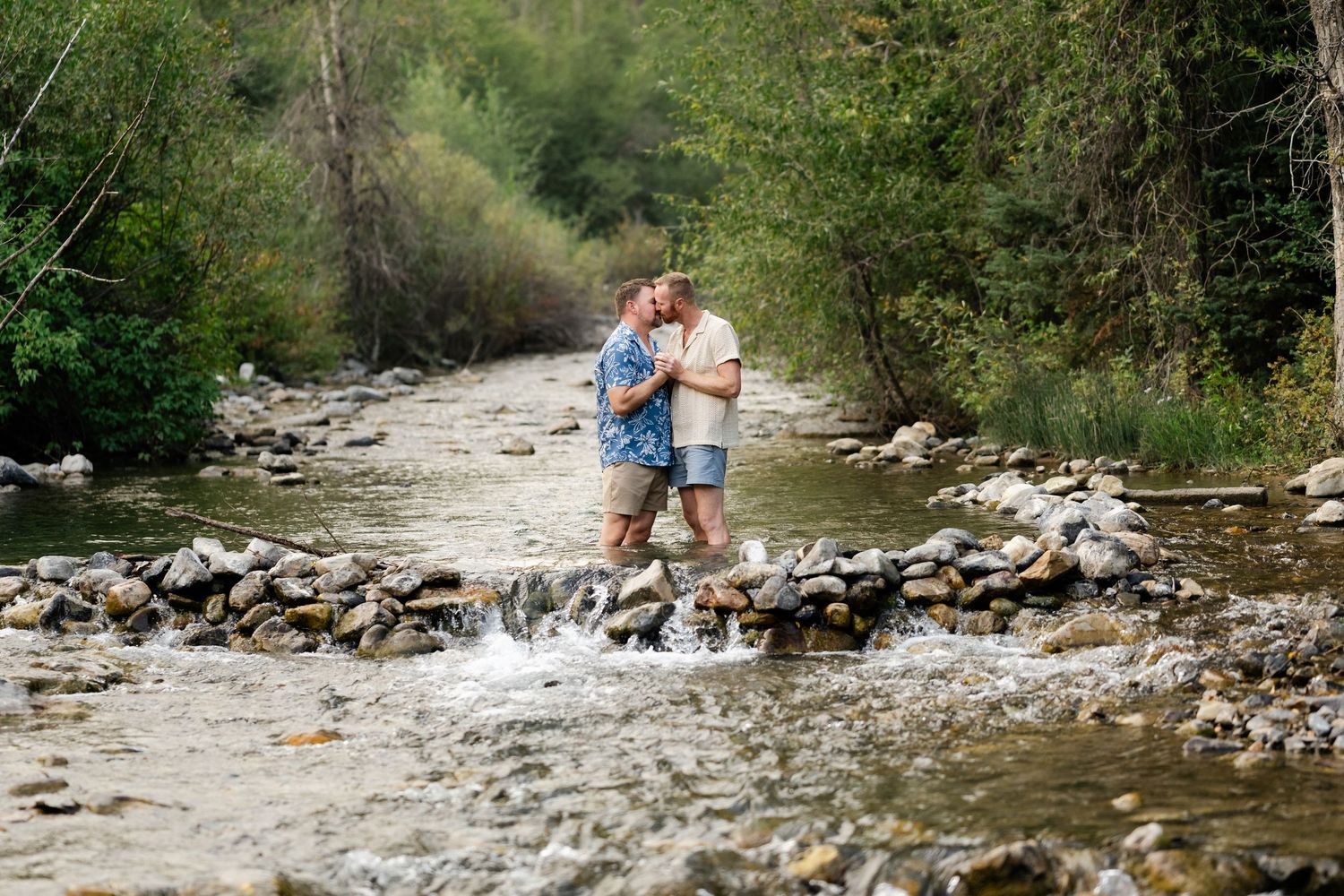 LGBTQ Couple kissing romantically while standing in a rocky mountain stream surrounded by lush green trees. Gay couple engagement photos.