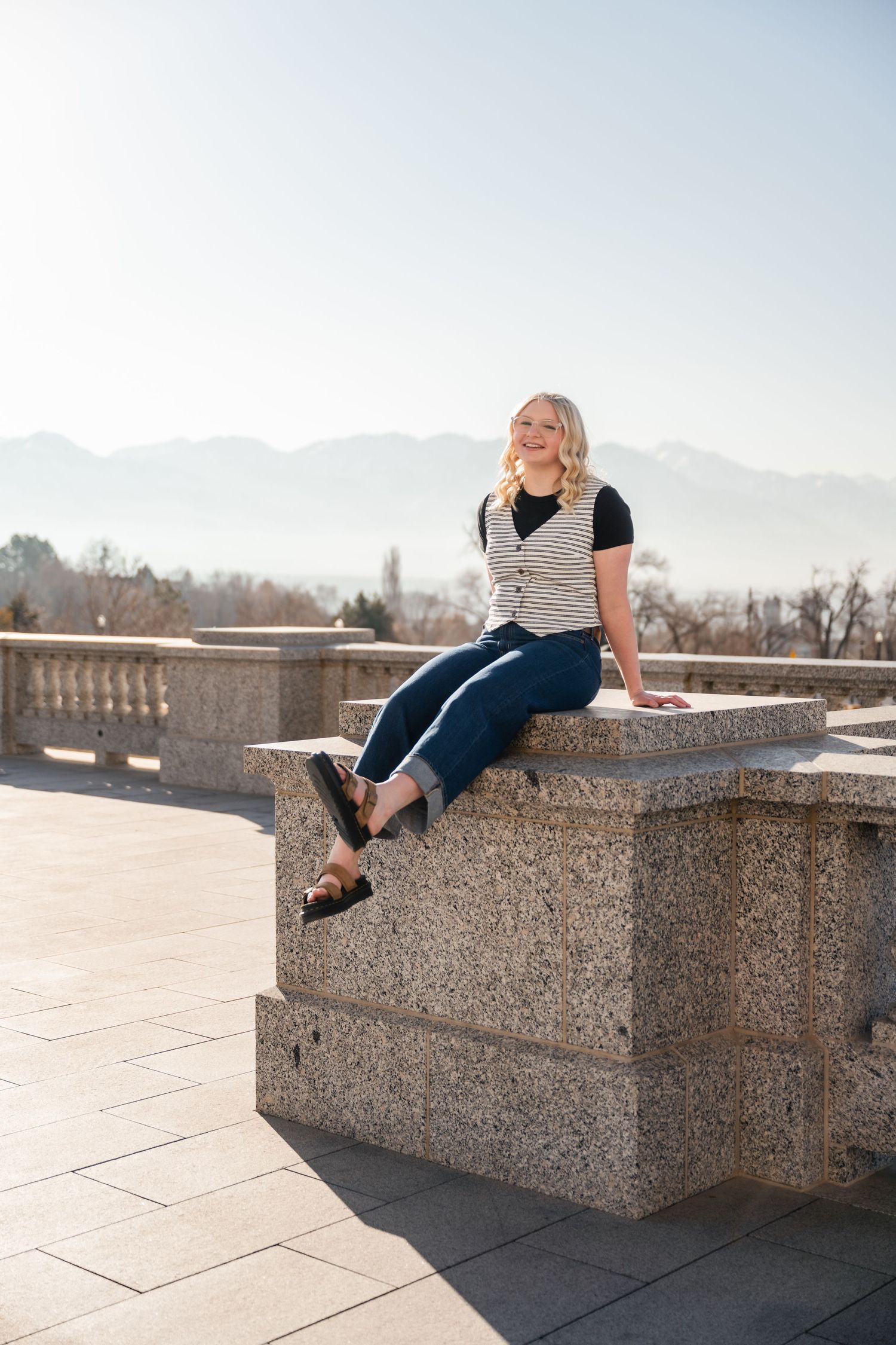 Someone sits casually on a stone ledge overlooking mountains on a sunny day wearing a striped vest and dark pants.
