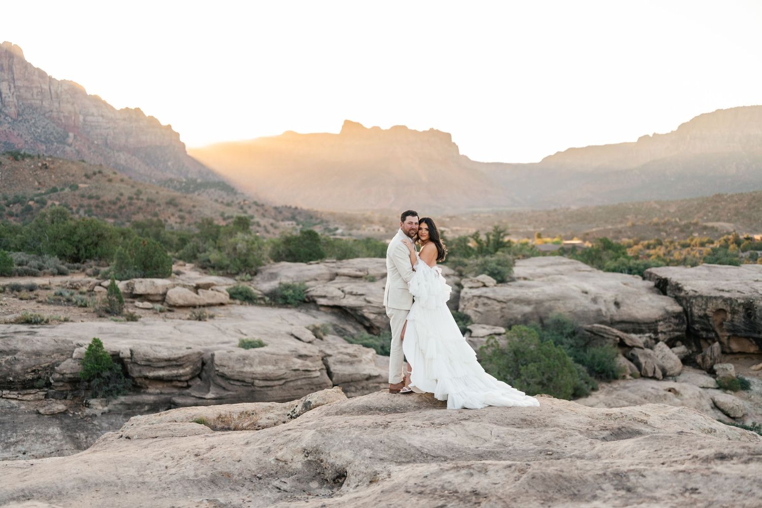 Couple embraces on scenic desert cliffs at sunset with dramatic mountain backdrop. Zion destination photographer.