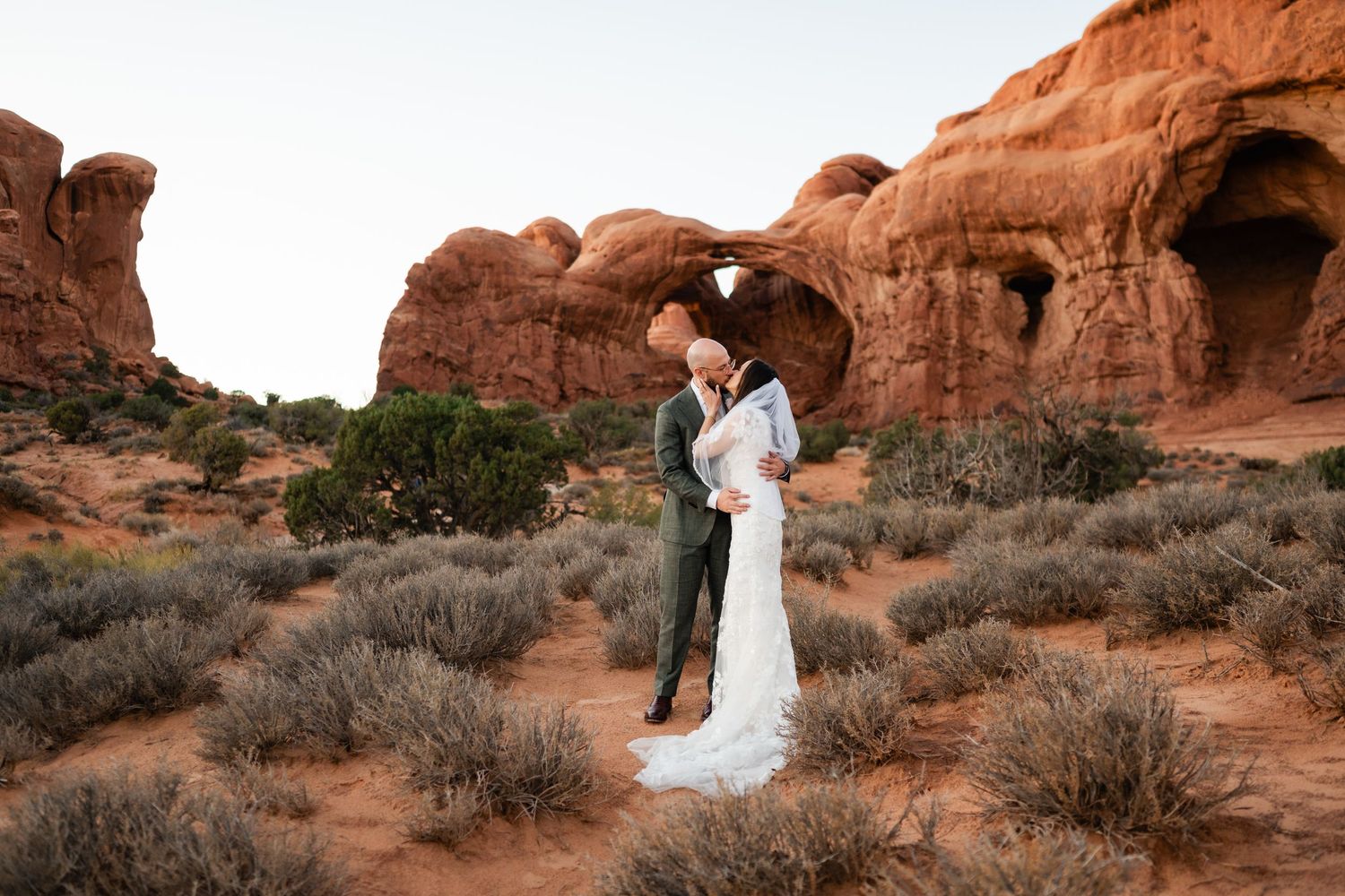 Couple in wedding attire stands against stunning red rock formations in Arches National Park at sunset.
