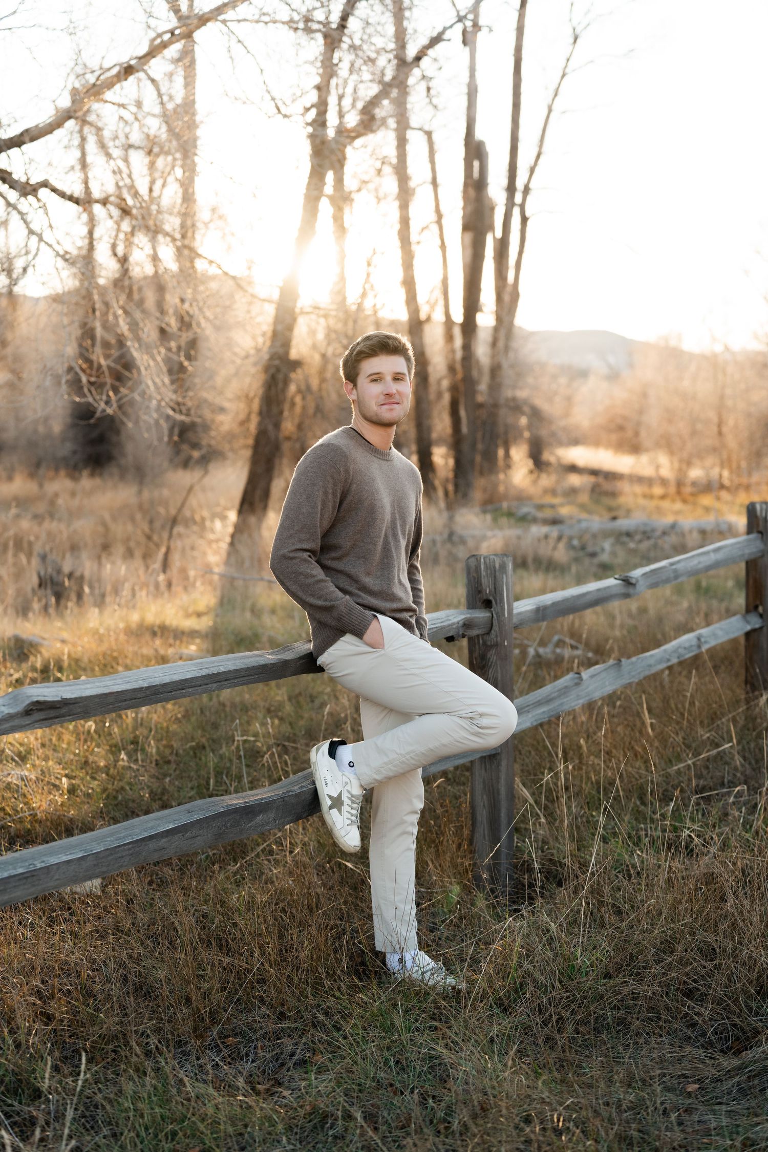 Person in casual attire sits casually on wooden fence rail during sunset with autumn foliage in background.