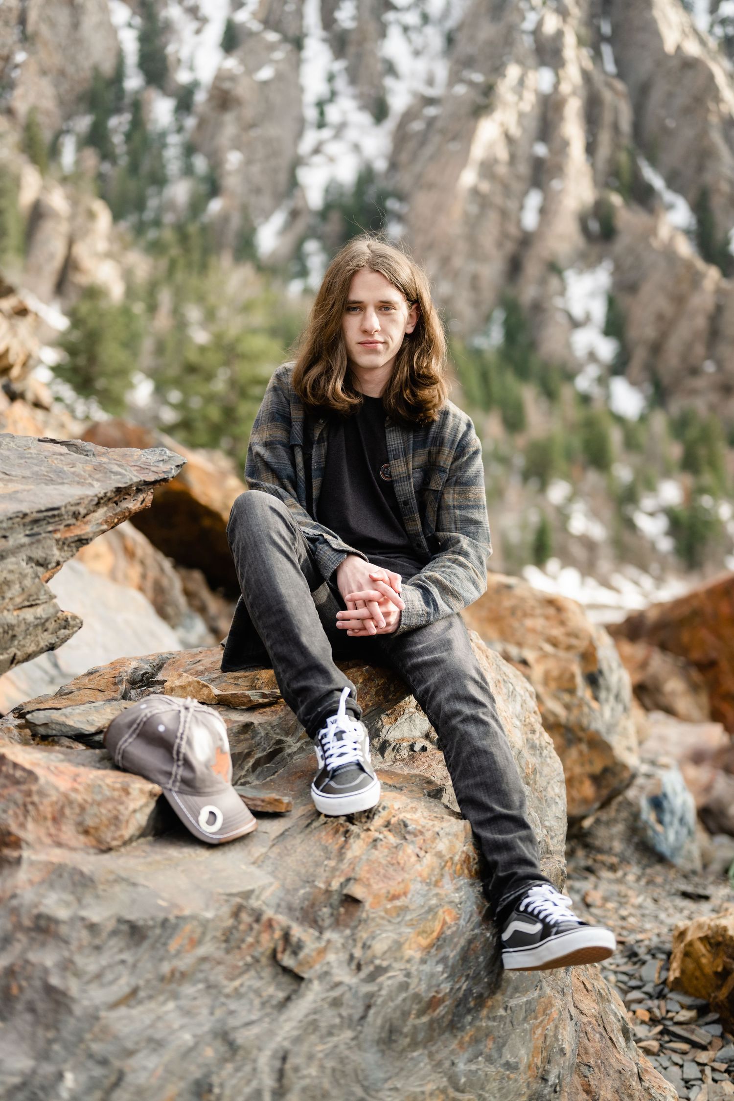 Person in black clothing and sneakers sits on rocky terrain in a natural outdoor setting with trees in background.