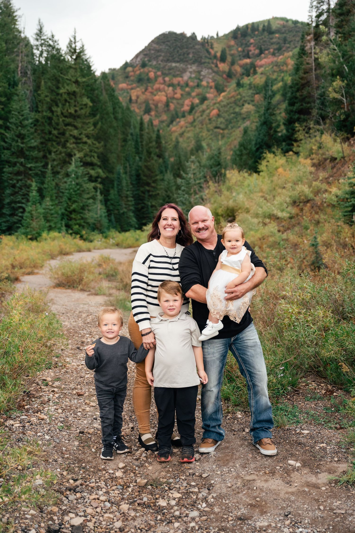 A family stands together on a hiking trail surrounded by evergreen trees and autumn foliage on a mountainside.