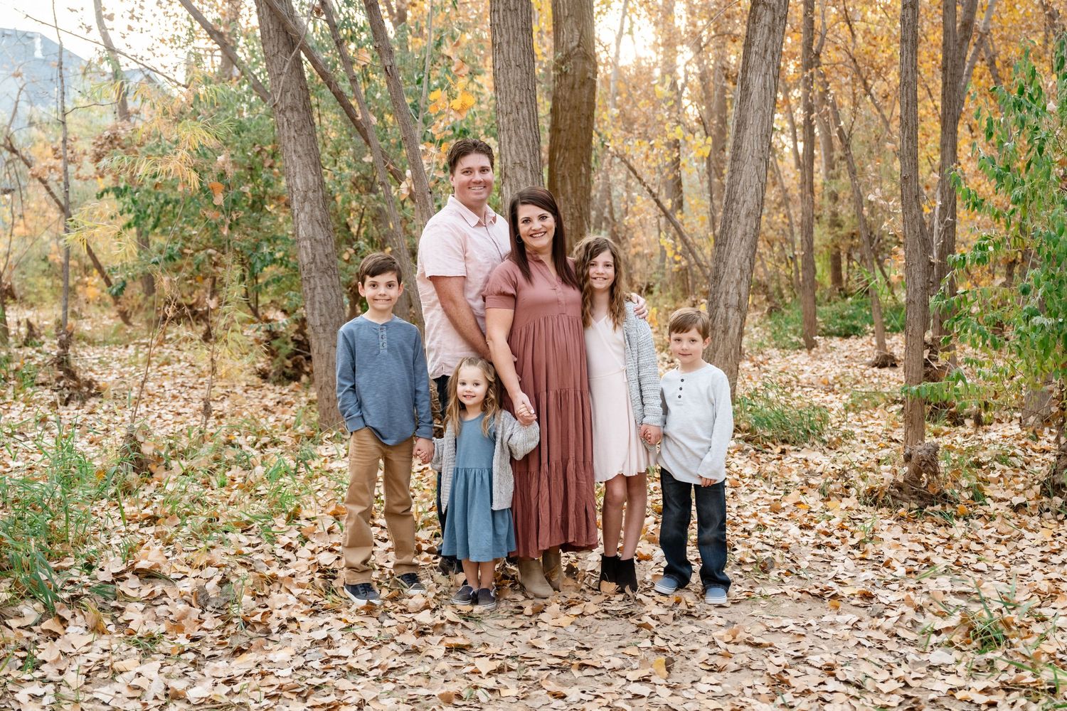 A family wearing coordinated neutral outfits poses together in a wooded area with fallen autumn leaves.