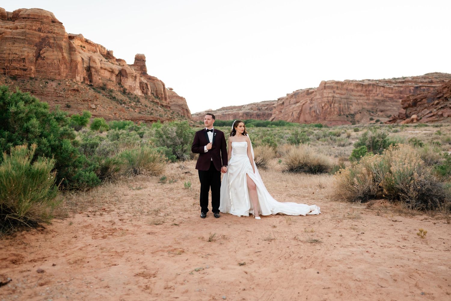 Bride and groom stand together in a stunning red rock desert canyon landscape during their outdoor wedding photo session.