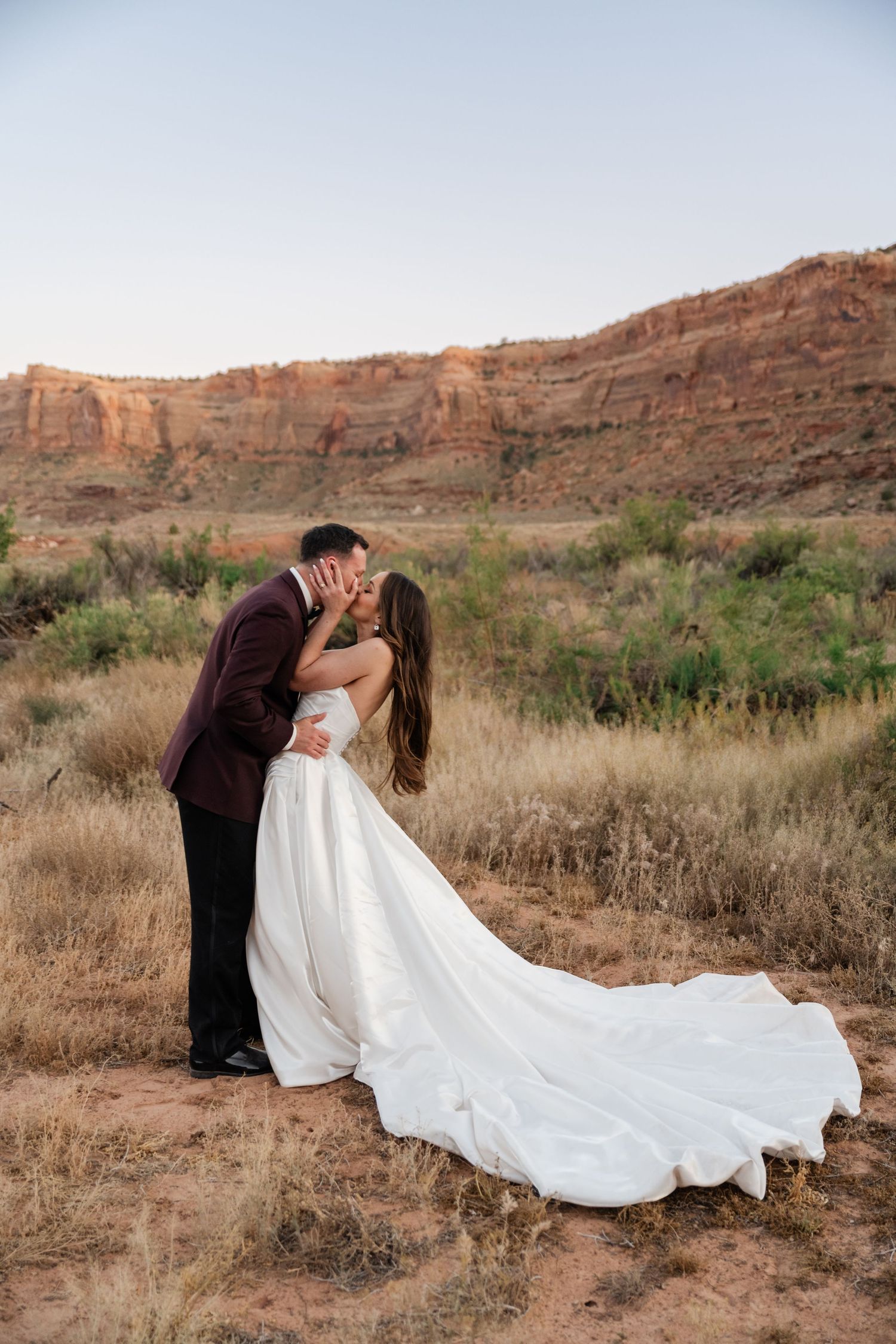 Bride and groom share a romantic kiss in a stunning desert landscape with red rock cliffs and golden grasslands at sunset.