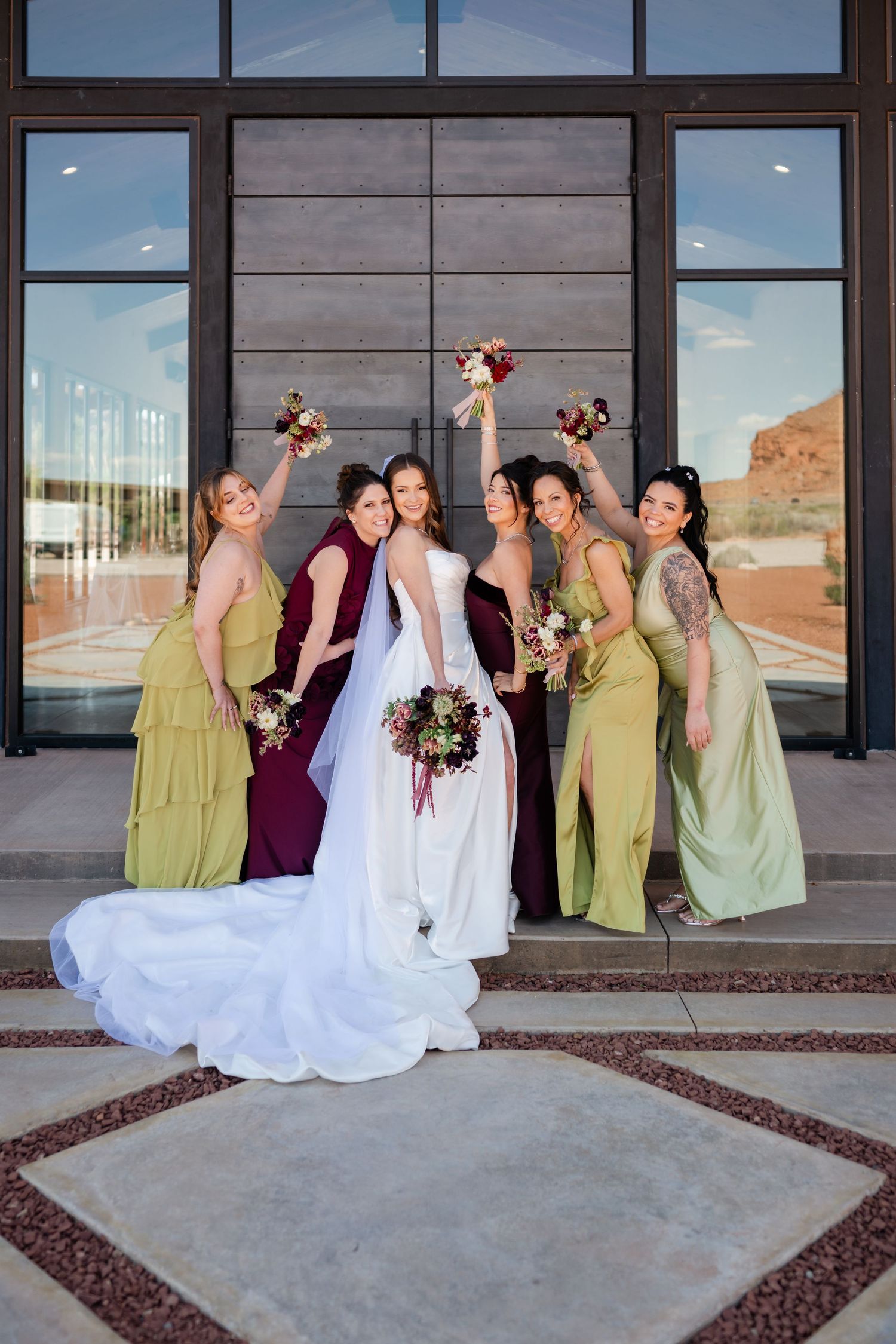 Bride in white gown poses with five bridesmaids in green and burgundy dresses, raising bouquets outside a modern venue.