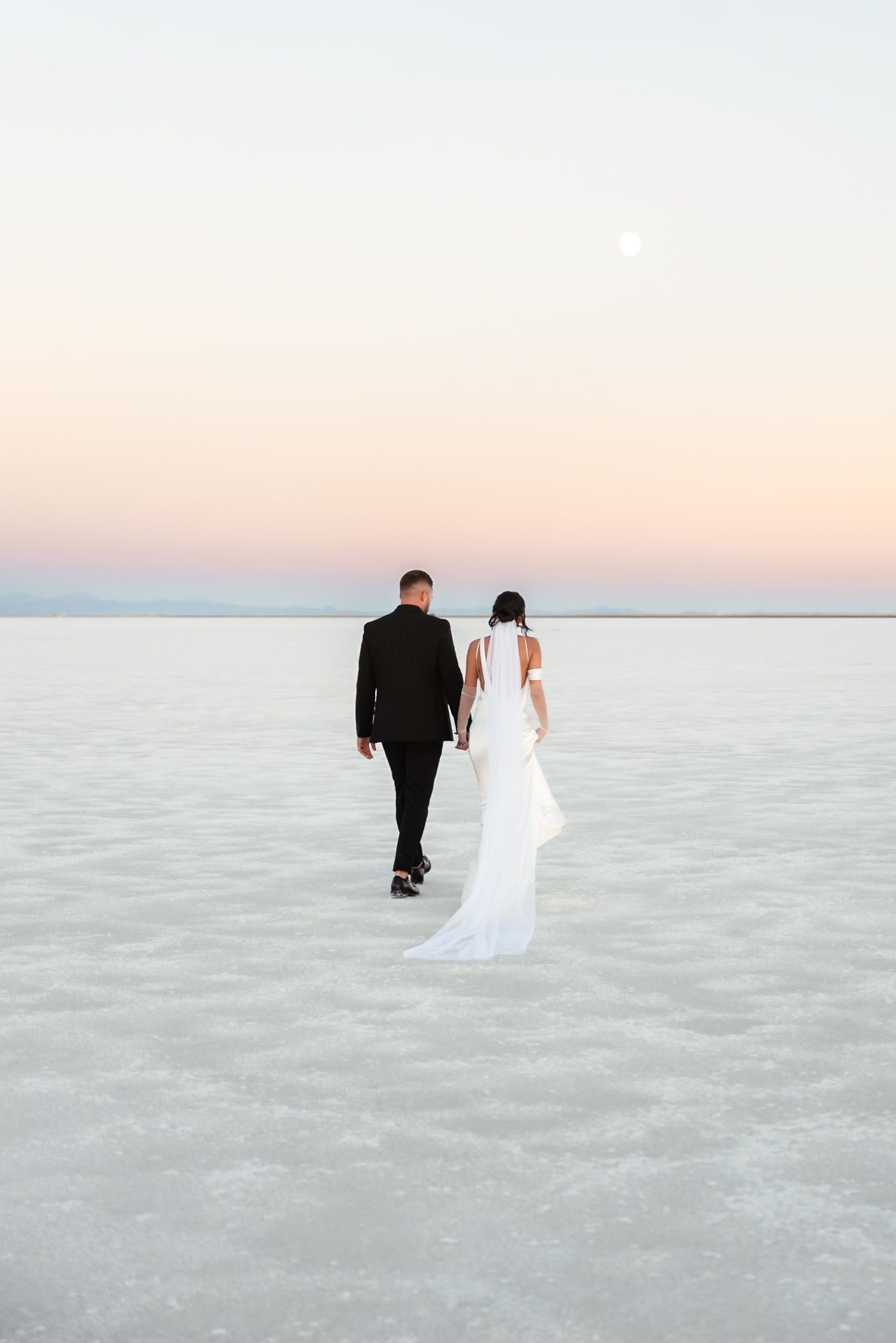 Couple walks hand in hand across ethereal salt flats during sunset.