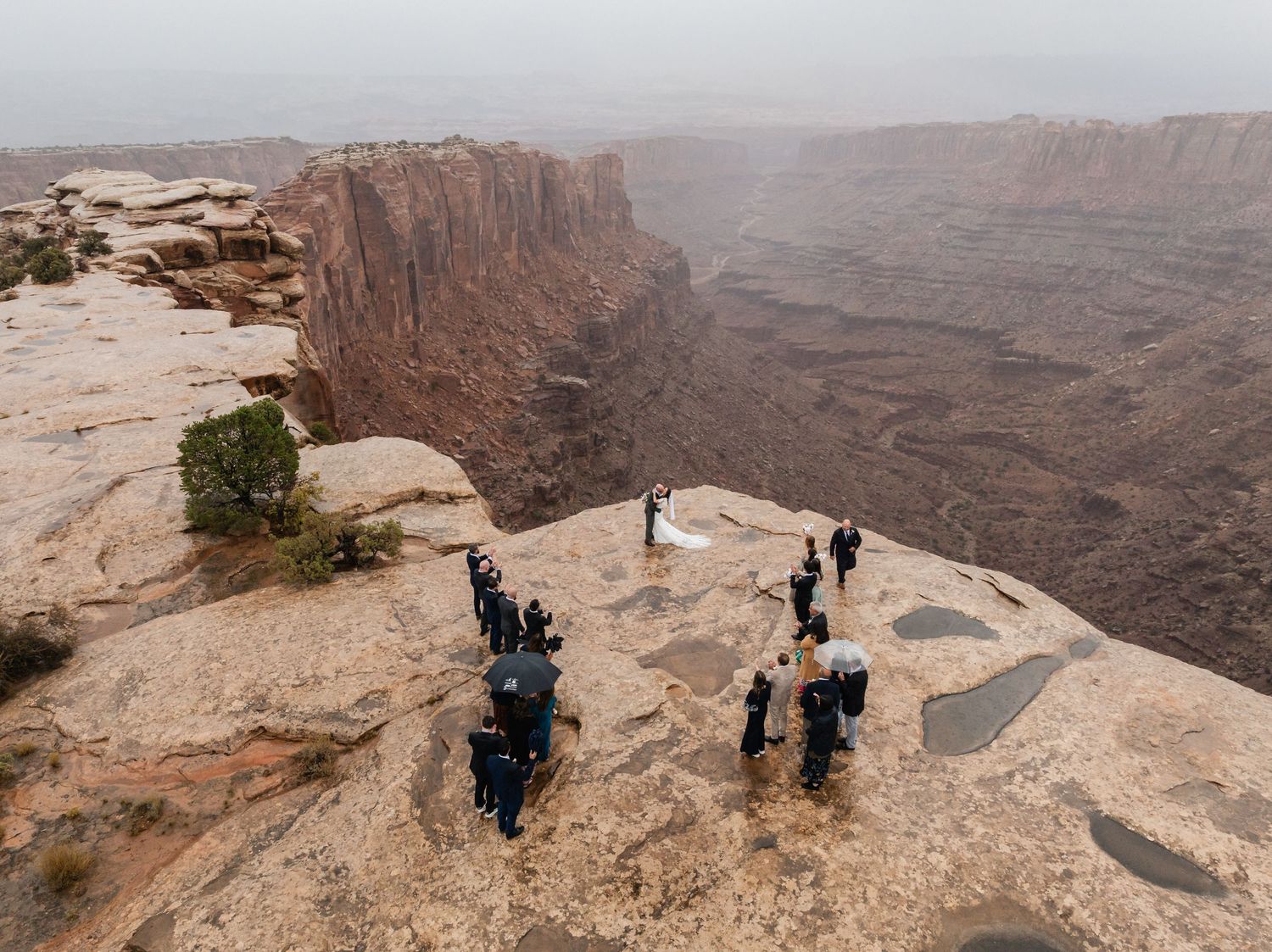 Wedding guests gather at the edge of a steep cliff overlooking a vast desert canyon with dramatic red rock formations during a ceremony.