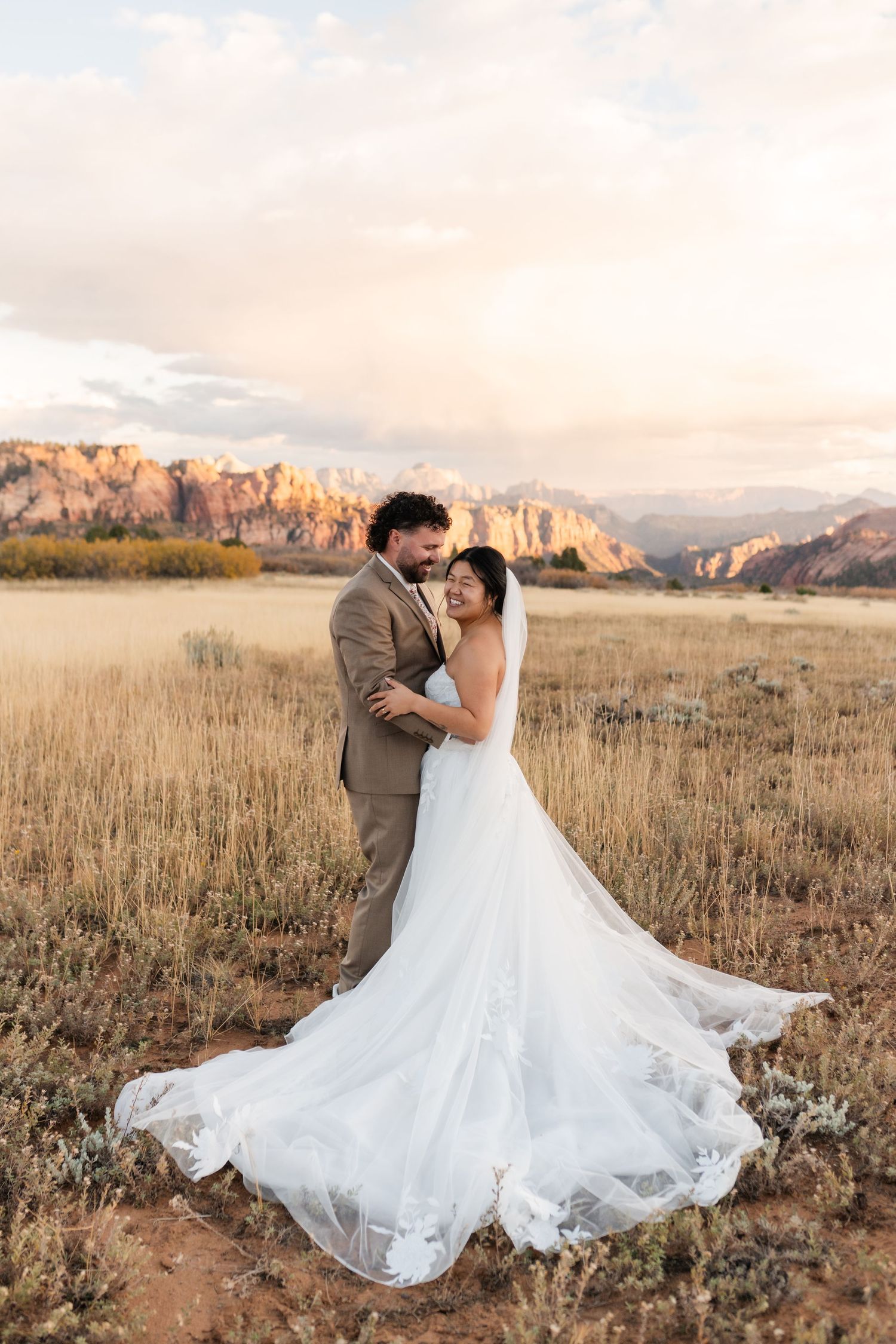 A wedding couple dances in a field with dramatic mountain cliffs and golden light in the background.