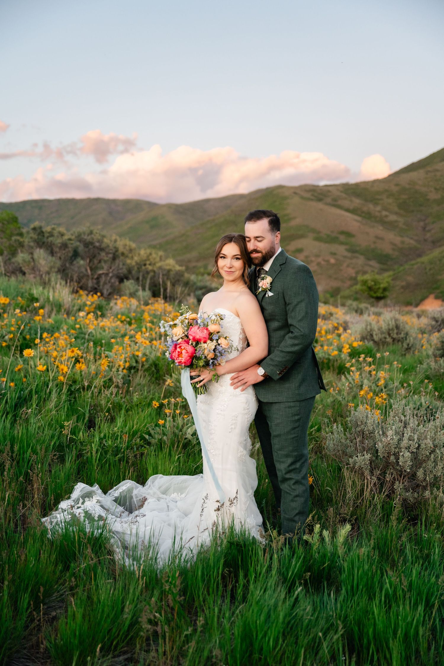 A wedding couple poses together in a field of yellow wildflowers at sunset with mountains in the background.