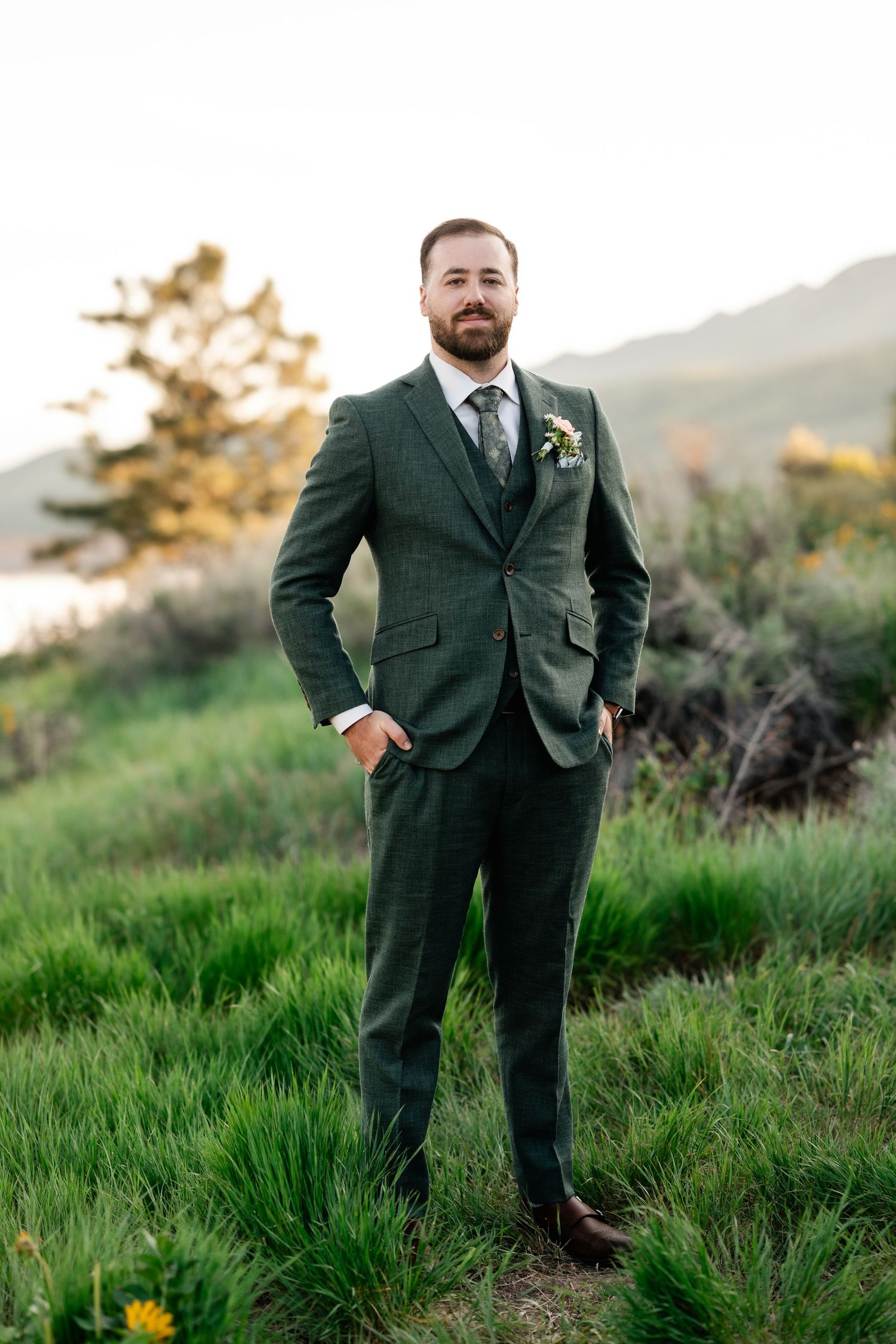 A groom in a dark green suit poses confidently in a grassy mountain landscape at sunset.