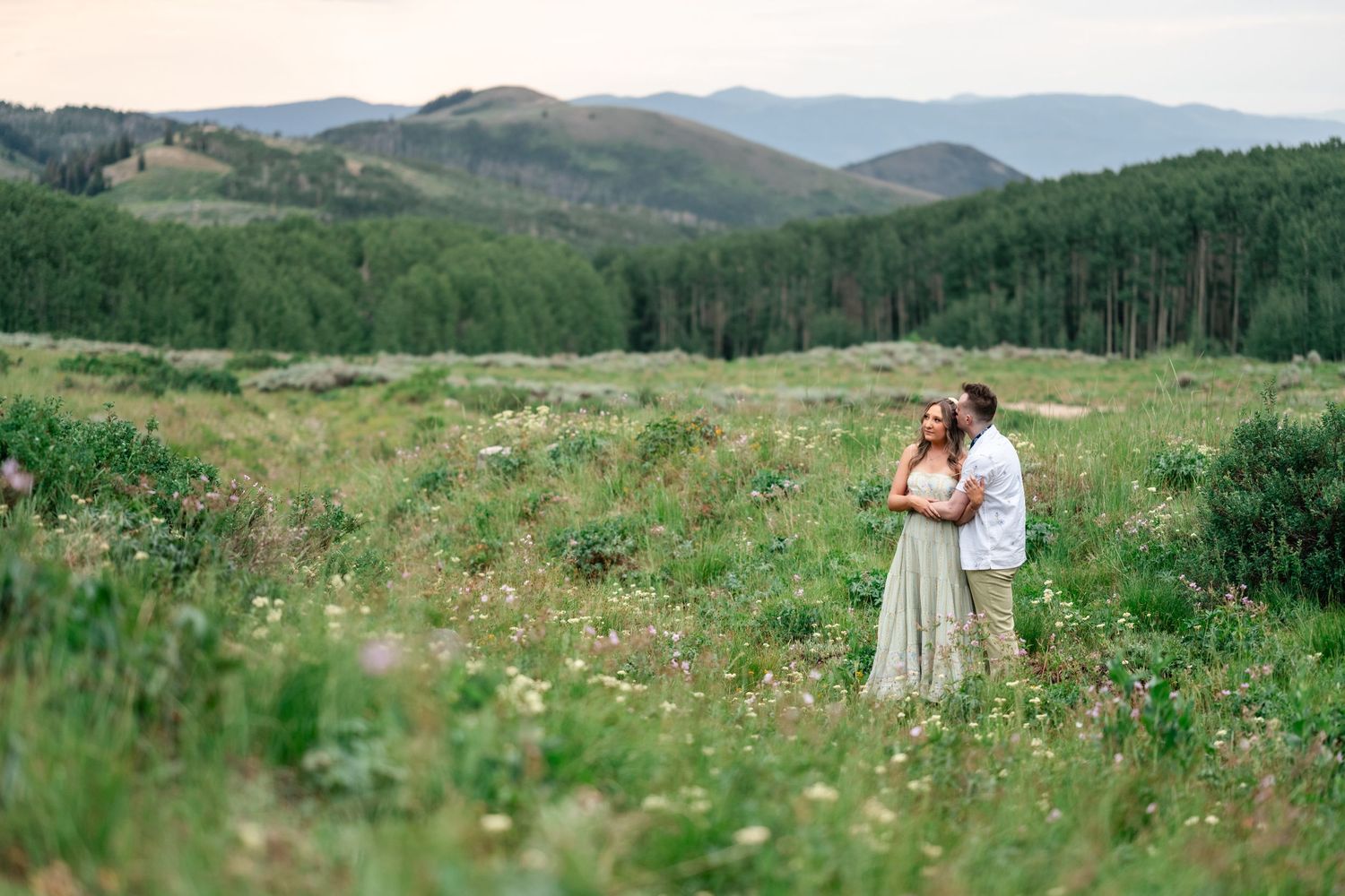 A couple embraces in a wildflower meadow surrounded by green mountains and forest on a summer afternoon.