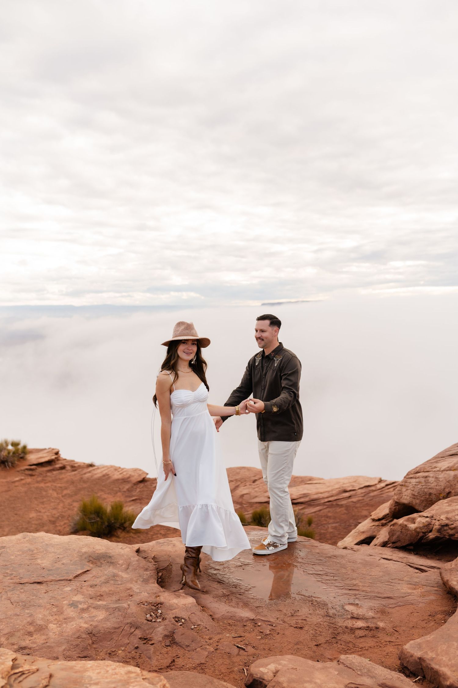 Couple in casual attire standing together on red rock cliff edge with cloudy sky backdrop.