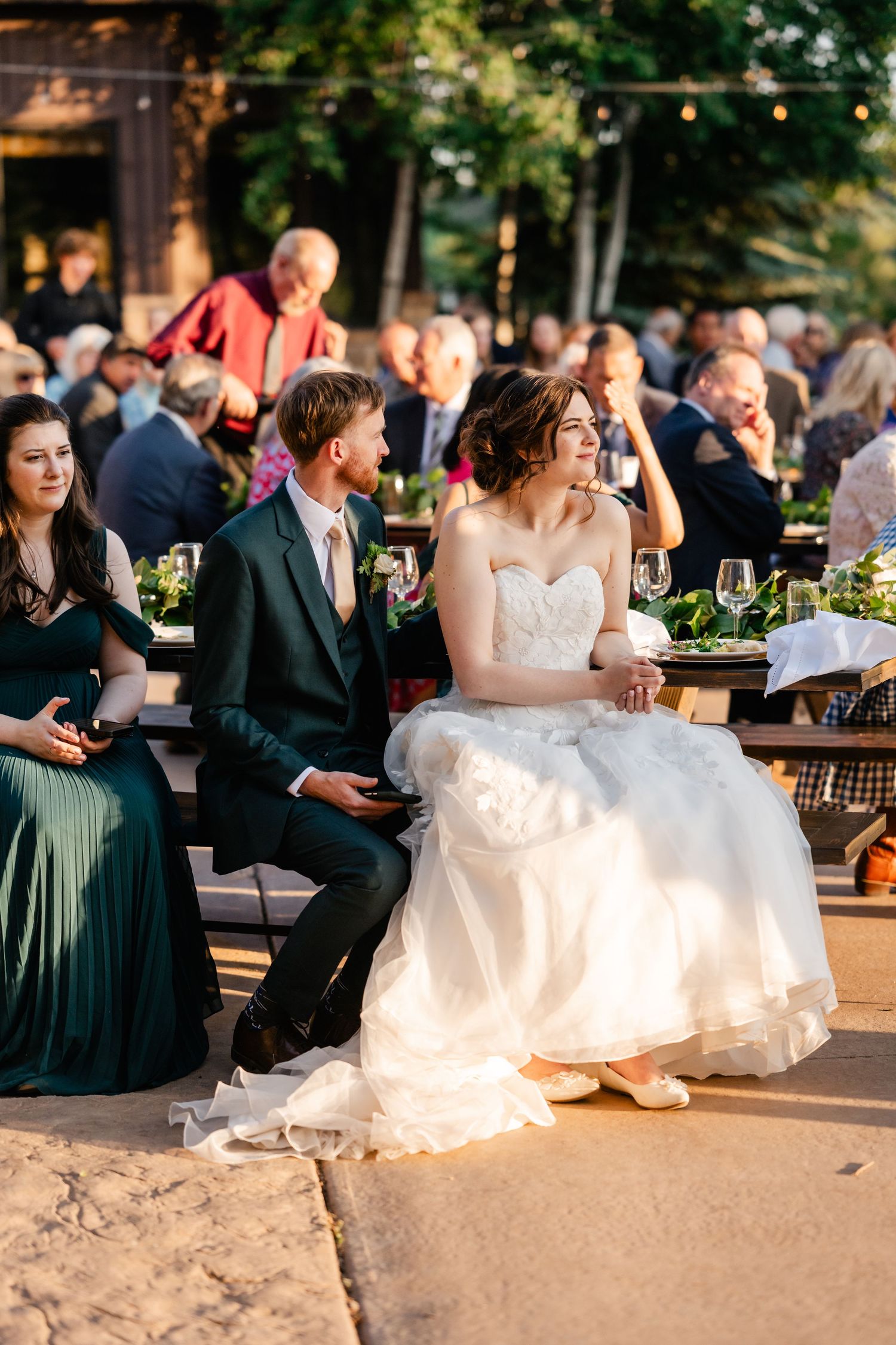 Wedding guests gather at an outdoor reception venue with string lights as a bride in white dress sits on steps at sunset.