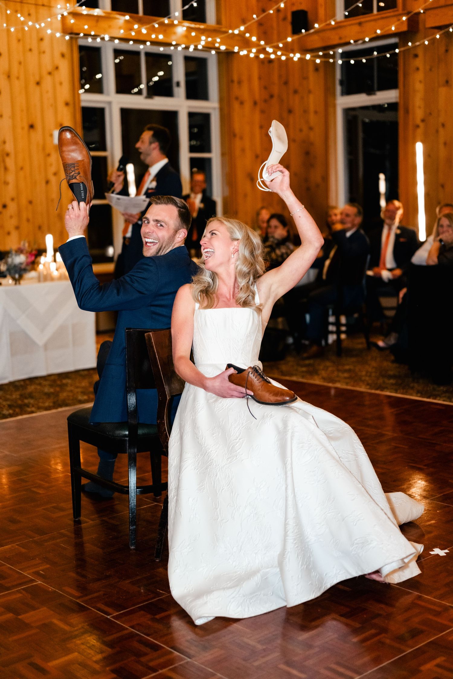 Couple dances energetically on a wooden dance floor under string lights at their reception.