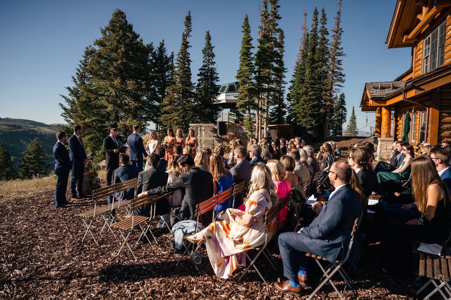Guests seated outdoors at a mountain wedding ceremony with evergreen trees and a rustic wooden lodge in the background.