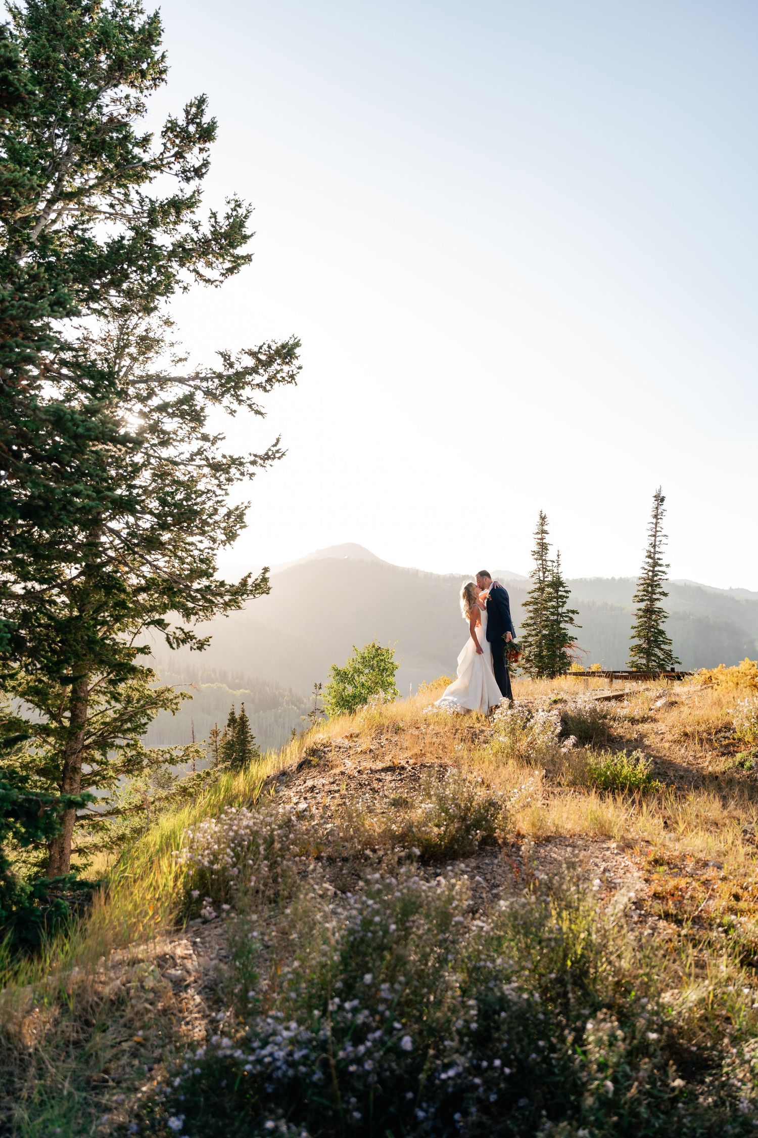 Couple standing on a scenic mountain ridge during golden hour with evergreen trees and mountains in the background.
