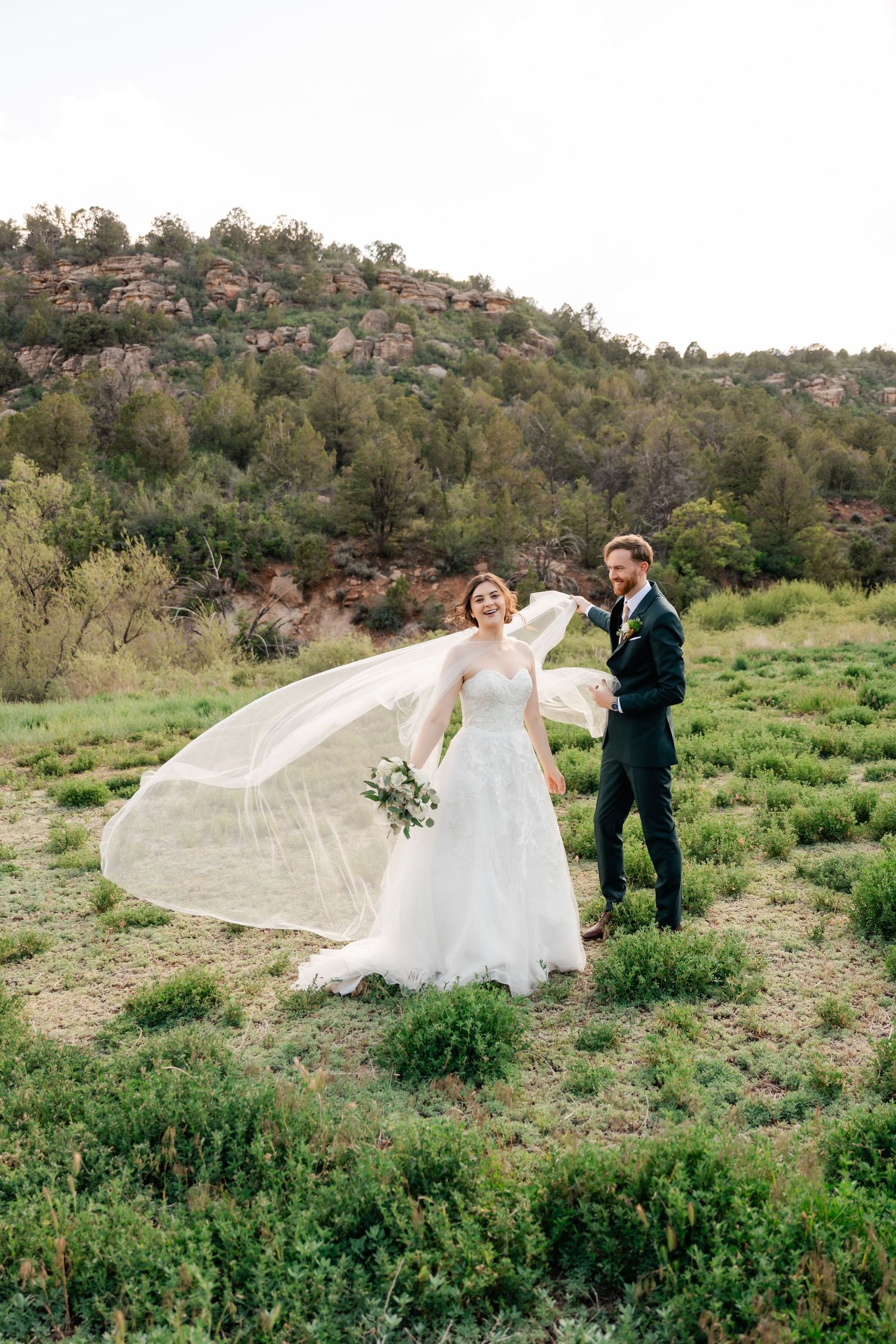 A wedding couple creates dramatic photos with a flowing veil against rocky mountain scenery.