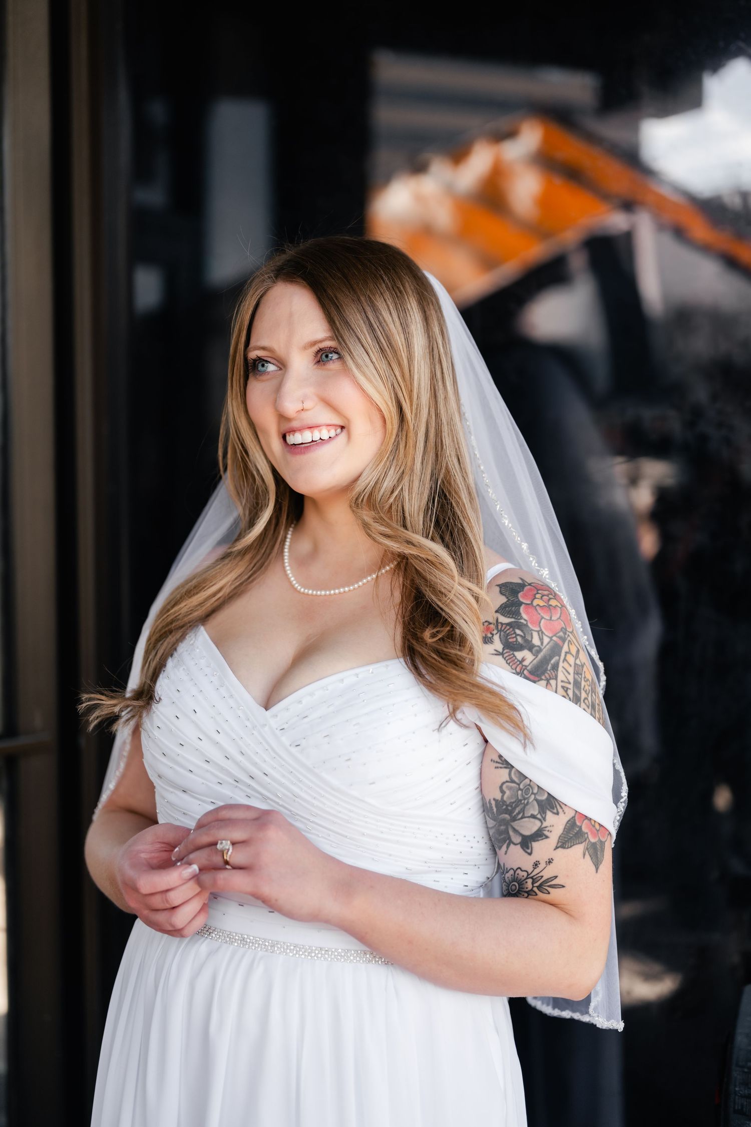 A bride in a white dress with visible tattoos smiles while wearing a flowing veil.