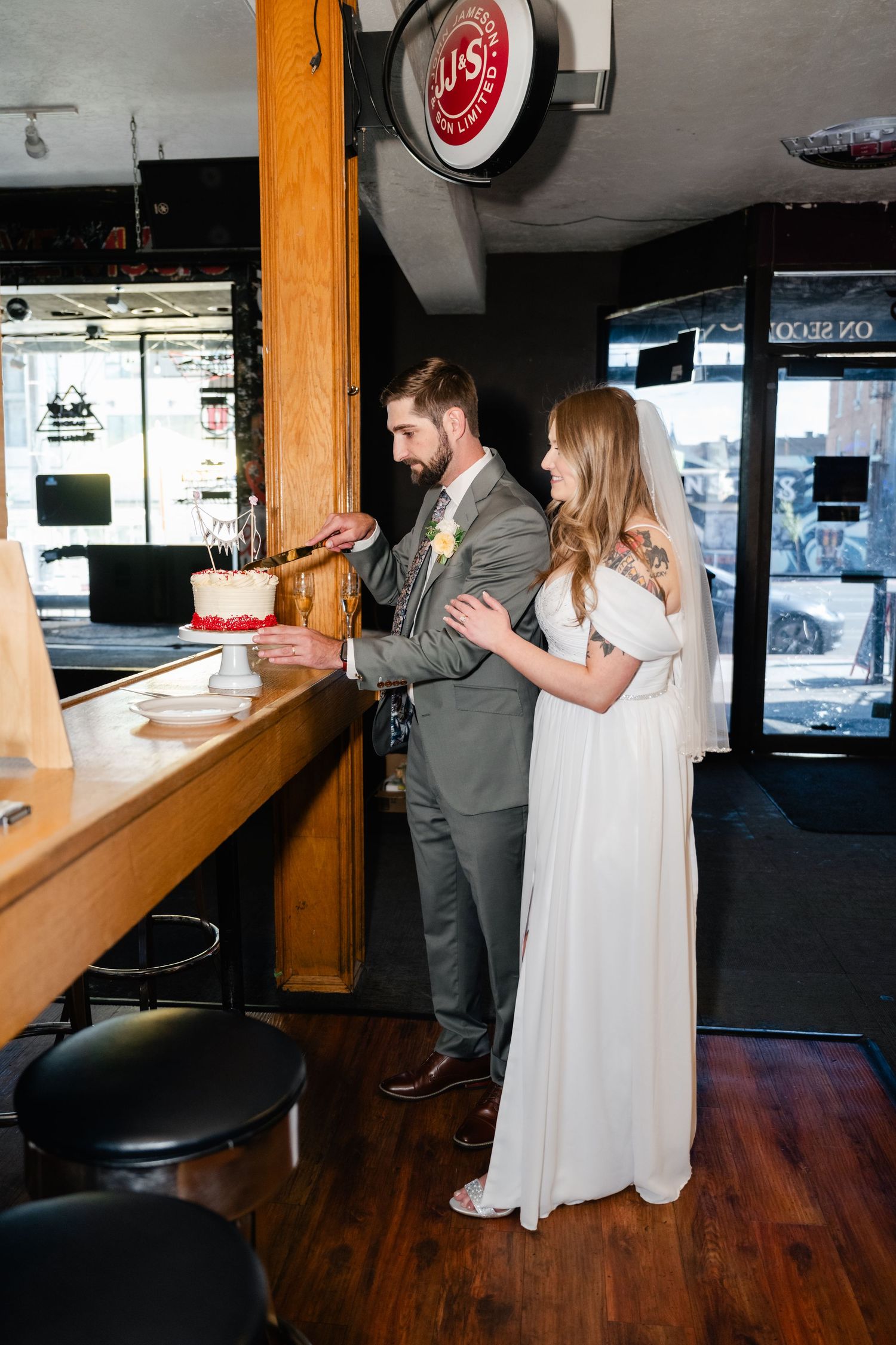 A couple in wedding attire cuts their wedding cake at a casual bar setting.