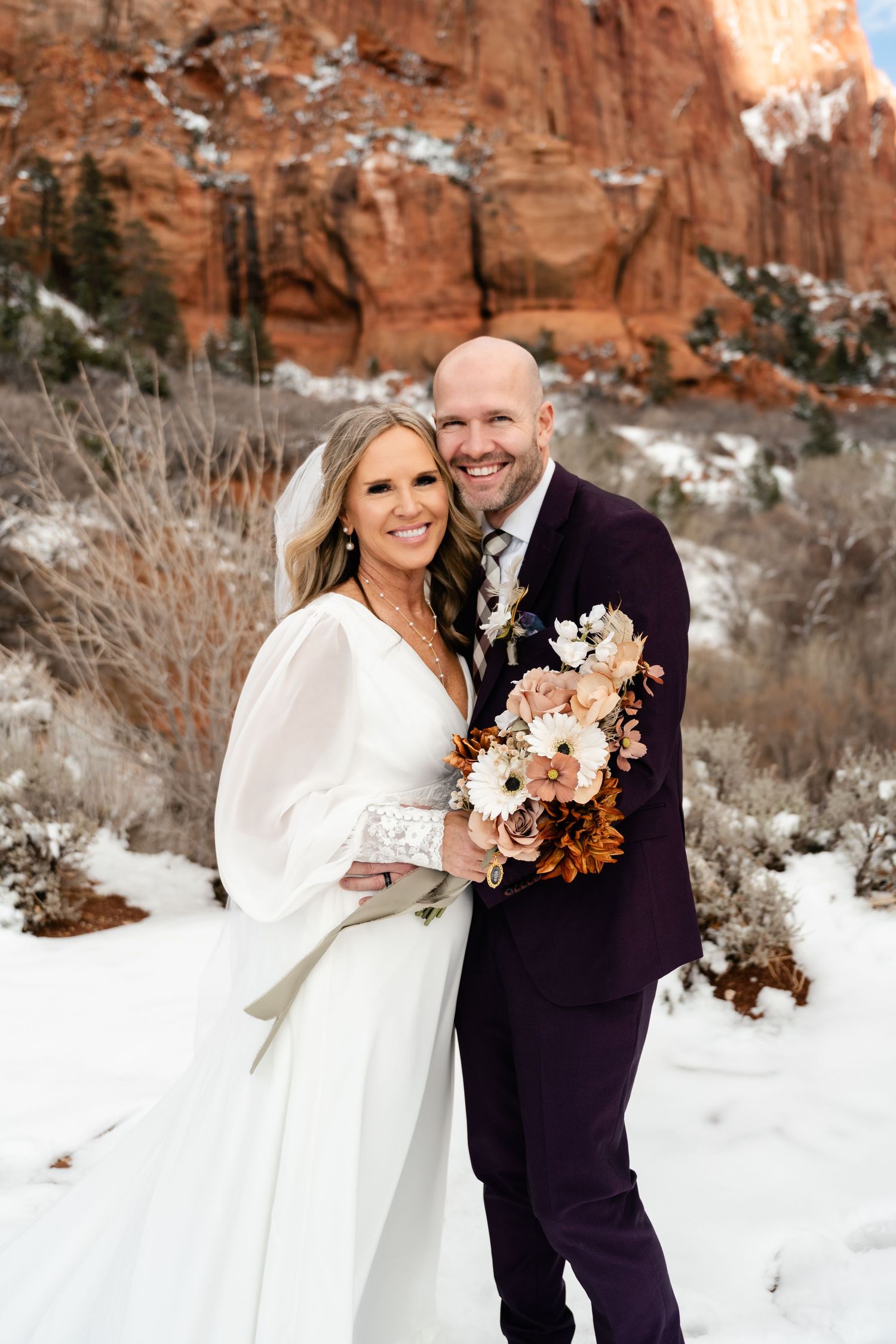 Winter wedding ceremony with white dress and burgundy suit against snowy red rock cliffs.