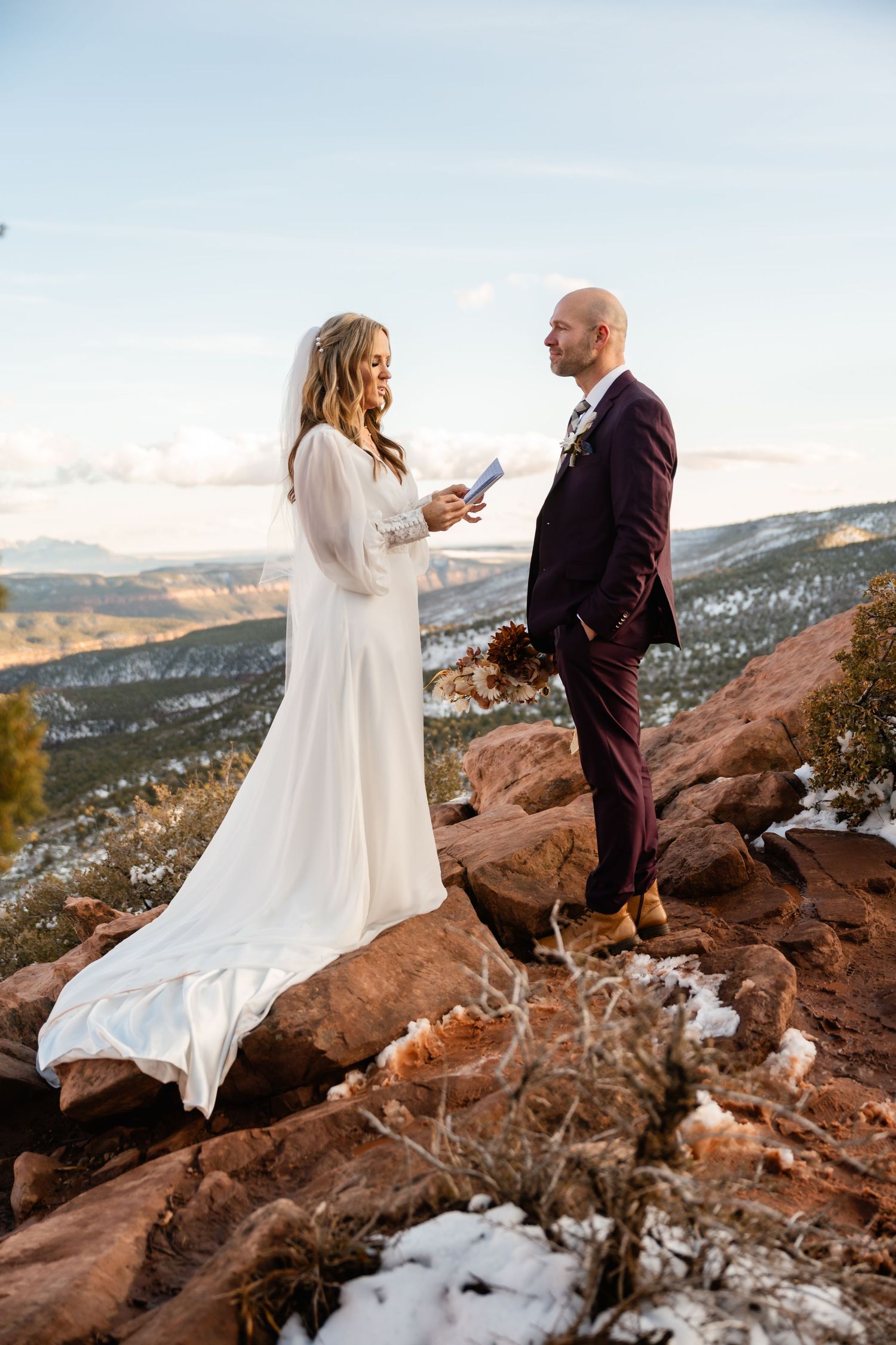 Couple in white dress and burgundy suit share a moment on snowy red rocks at sunset.