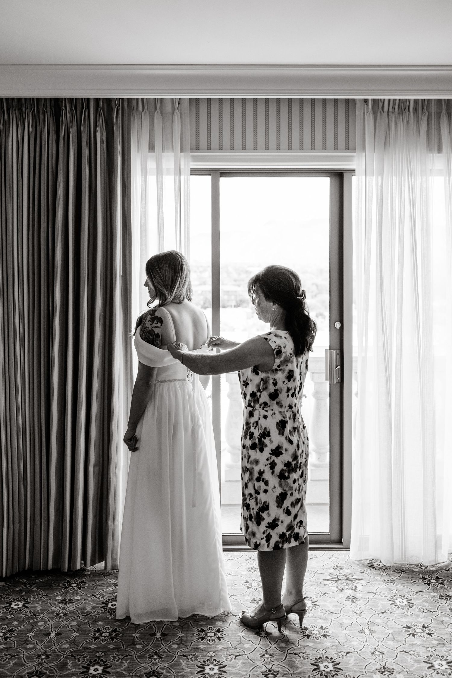 Black and white photo of a woman helping adjust a flowing white dress by a window with elegant curtains.
