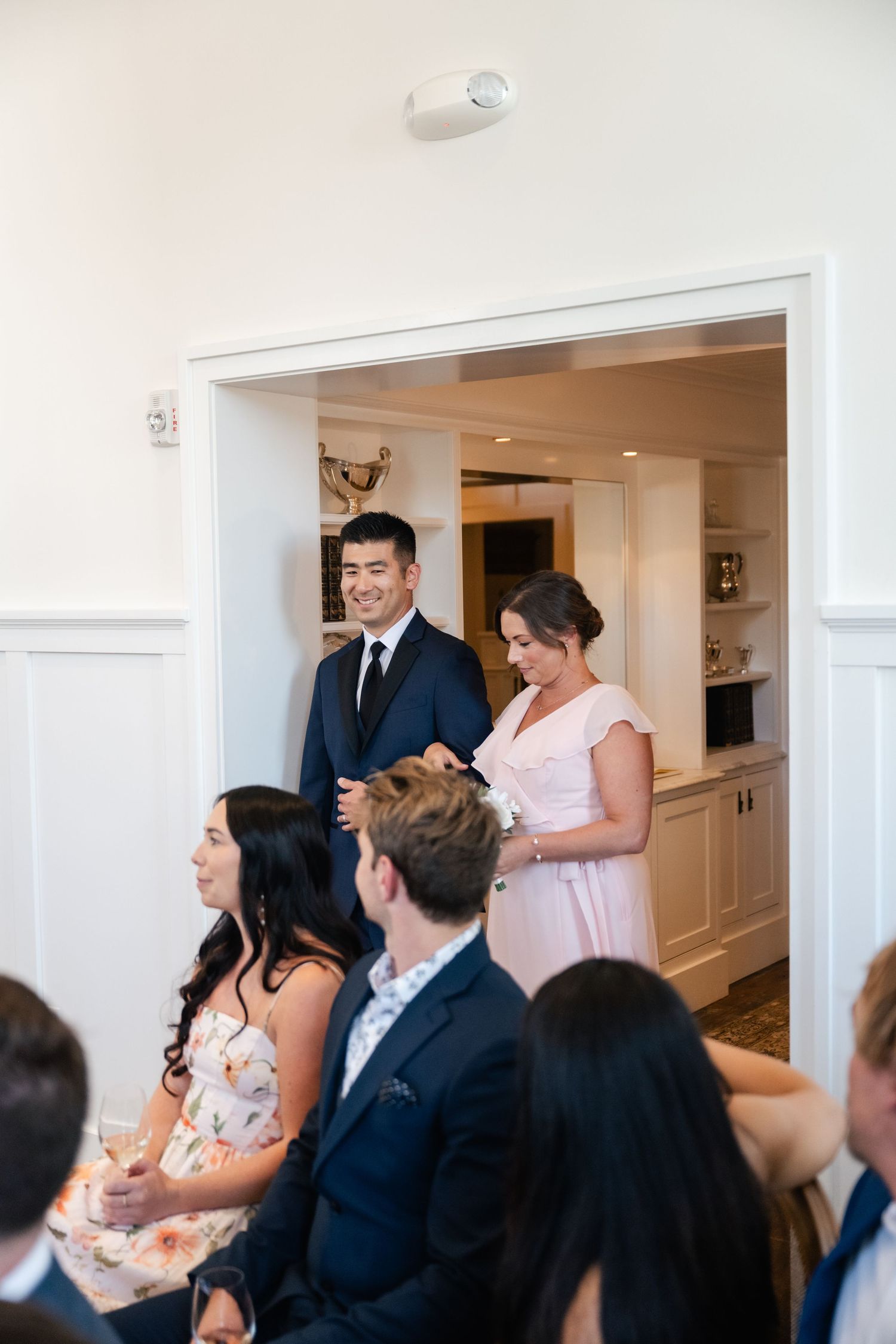 Wedding guests seated in a bright white room smile and laugh during a reception celebration.