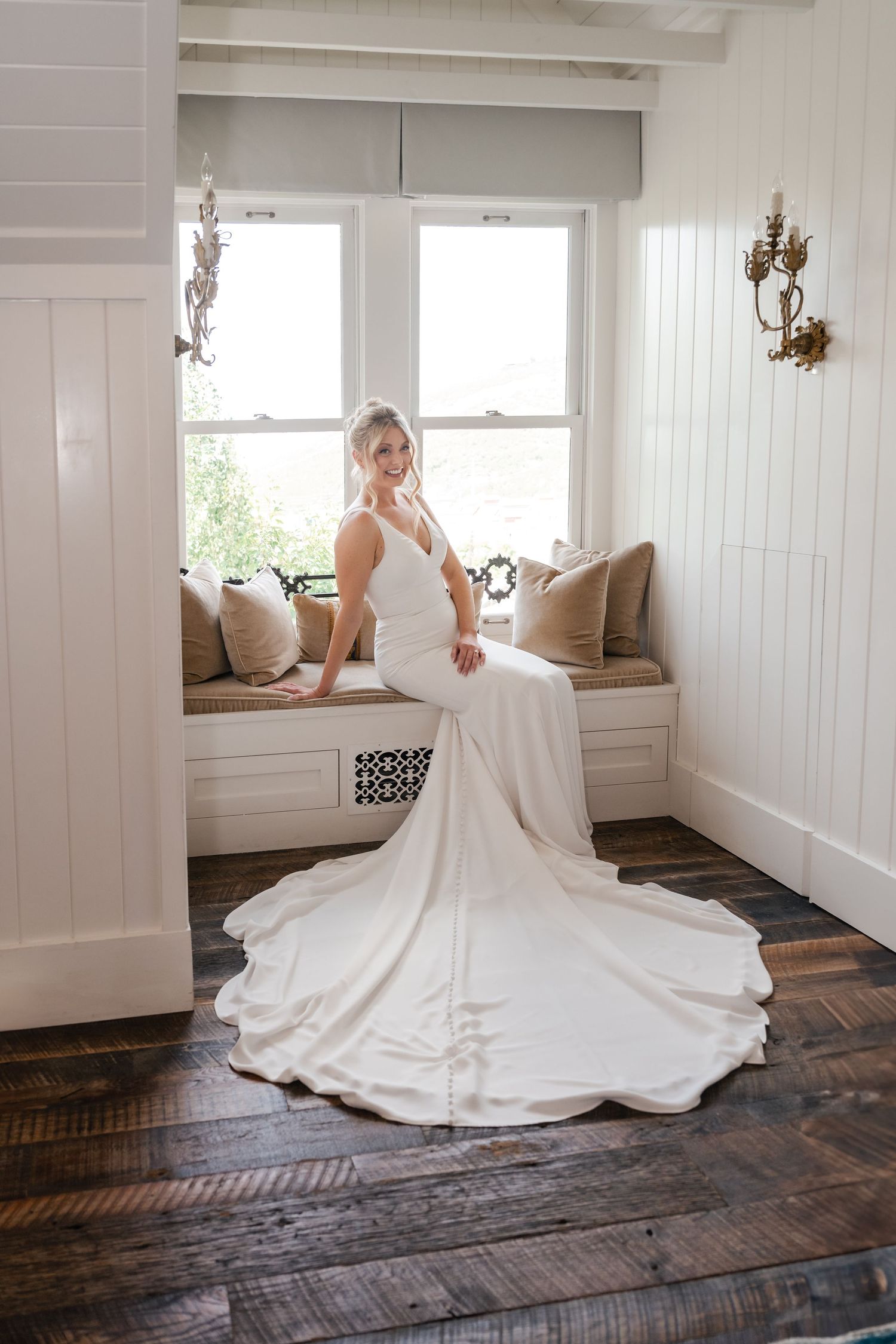 Elegant white wedding dress with long train spread across dark wooden floor in sunlit room.