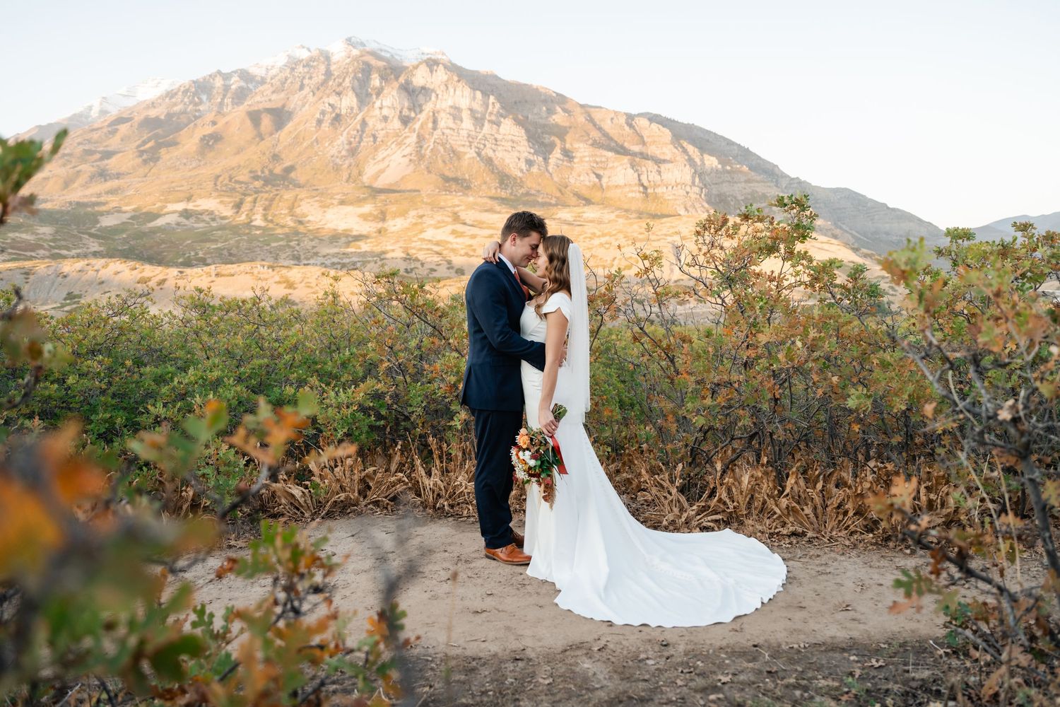 Couple embraces in front of majestic mountain landscape during golden hour wedding photo session. Best Utah Wedding Photographer.