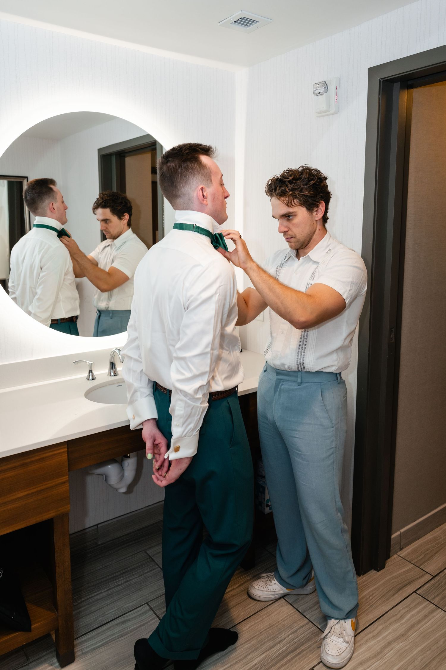 Two people in formal attire help each other get dressed in front of a bathroom mirror.
