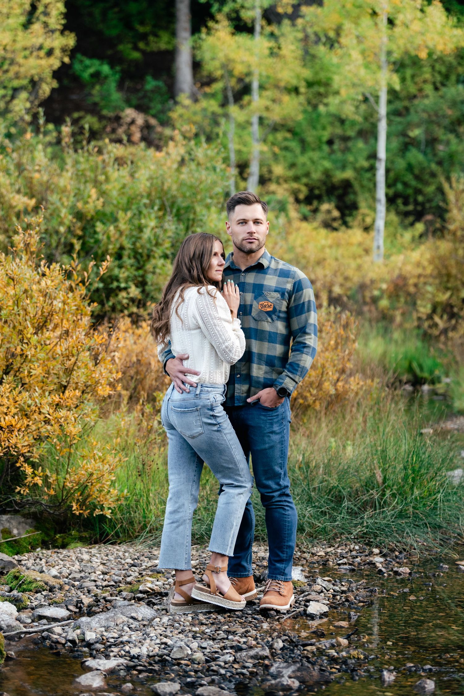 A couple stands together on rocky ground near a stream surrounded by autumn foliage.