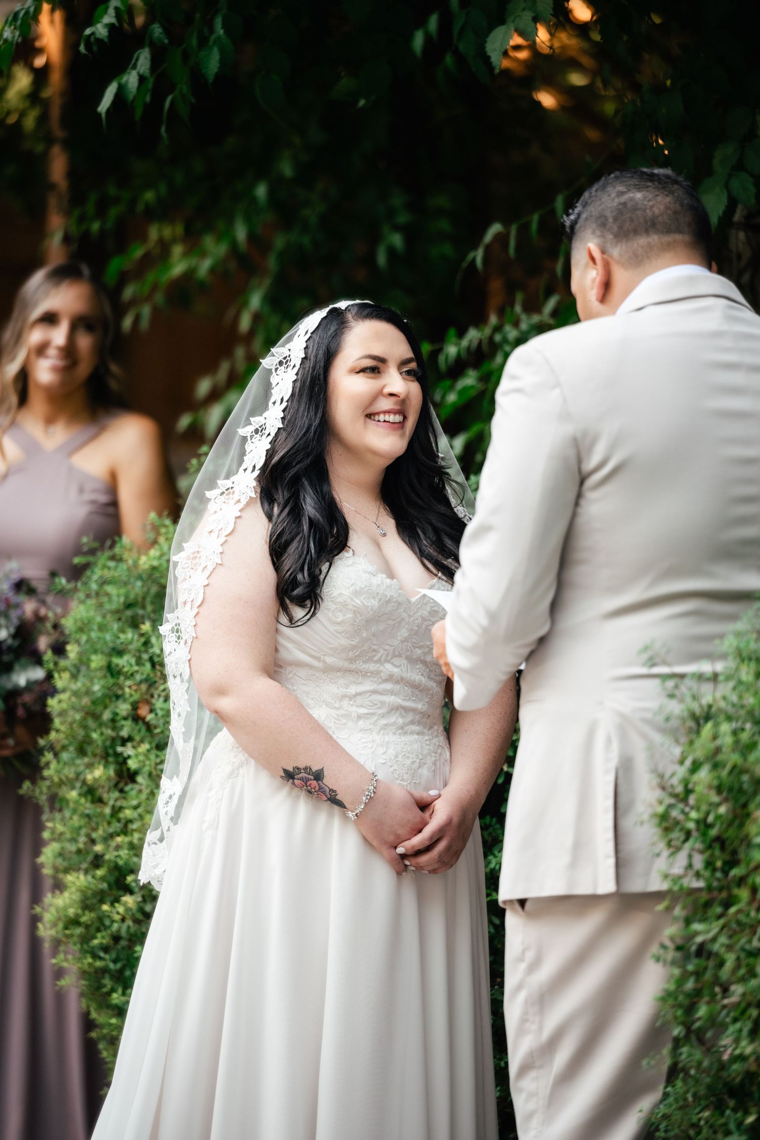 A joyful wedding ceremony takes place outdoors with a couple exchanging vows under green foliage and string lights.