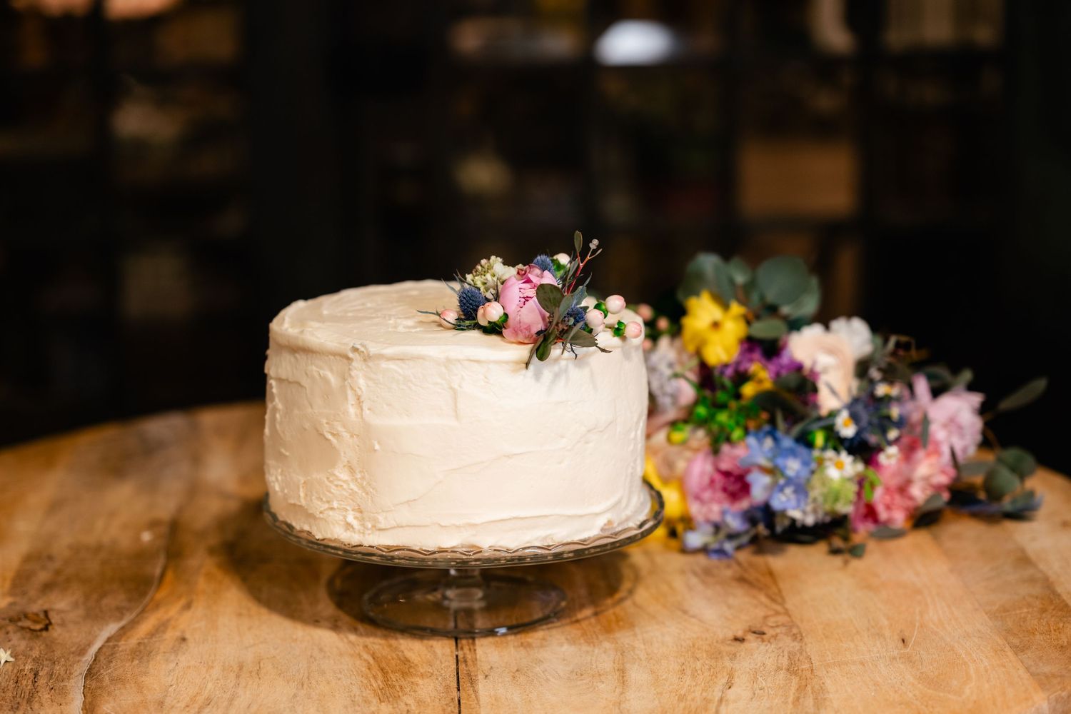 White frosted layer cake decorated with fresh colorful flowers on wooden table with rustic styling.